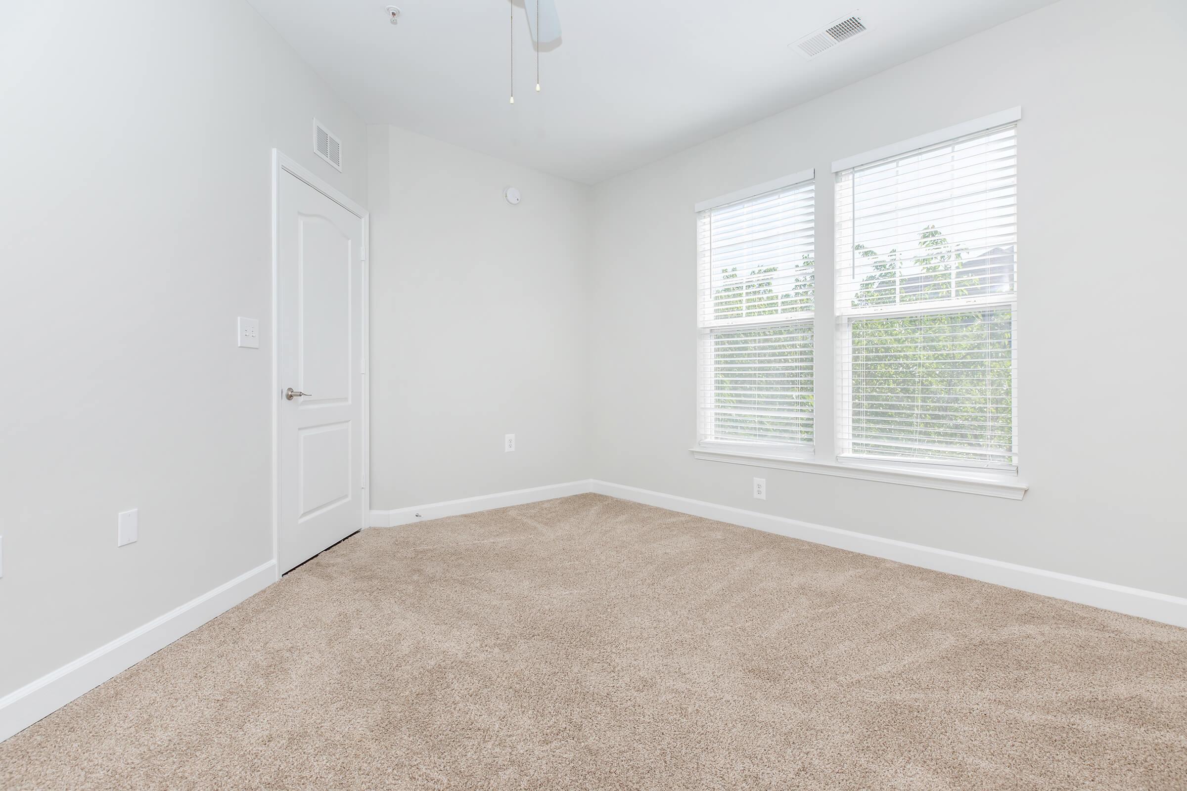 A bright, empty room with light gray walls and beige carpet. It features two large windows with white blinds, allowing natural light to flood in. A white door is visible on the left side, and the ceiling has a ceiling fan. The overall space appears clean and well-maintained, suitable for various uses.