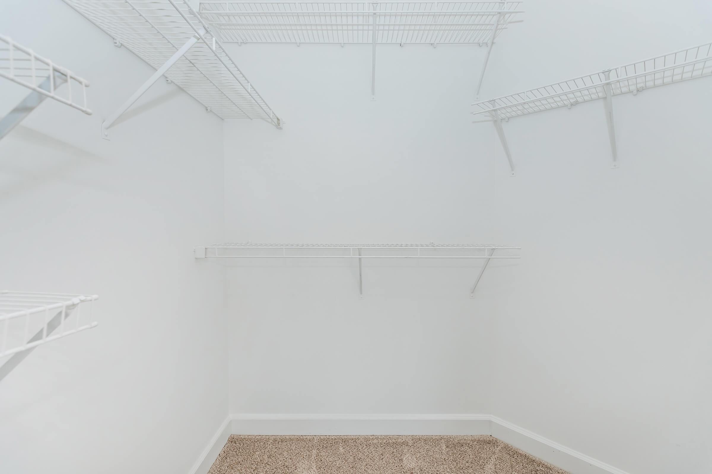 Empty walk-in closet with white walls and wire shelving. The floor is covered in beige carpet. There are several shelves mounted on the walls, creating an open and spacious storage area.