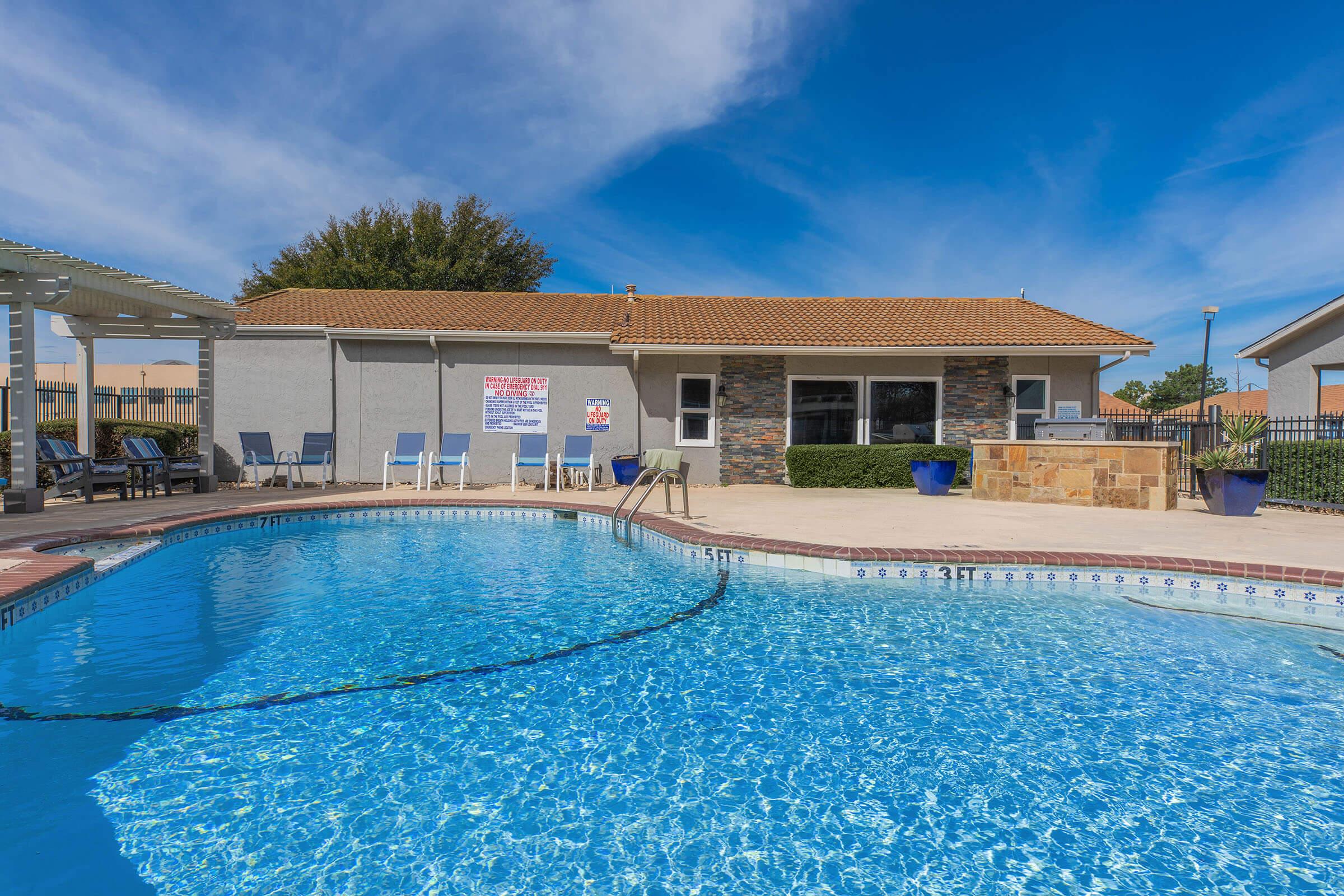 A bright outdoor swimming pool with clear blue water, surrounded by lounge chairs and potted plants. In the background, there is a building featuring a stone facade and a covered patio area. The sky is clear and sunny, contributing to a relaxed, inviting atmosphere.