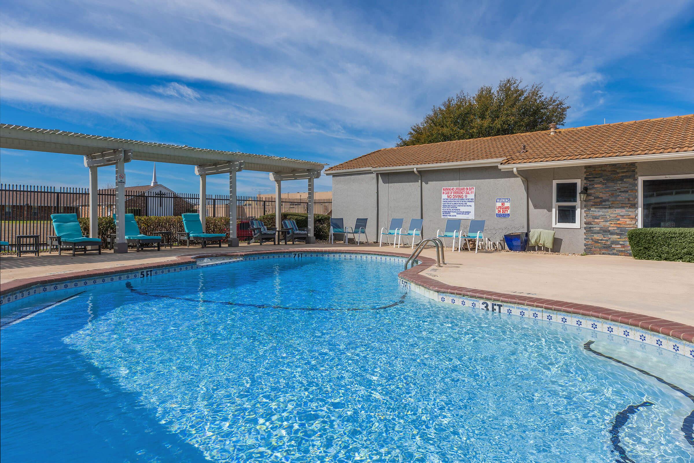 A clear blue swimming pool surrounded by lounge chairs and a shaded area, with a concrete deck. A building is visible in the background with a sign on it. The sky is partly cloudy, creating a bright and welcoming atmosphere.