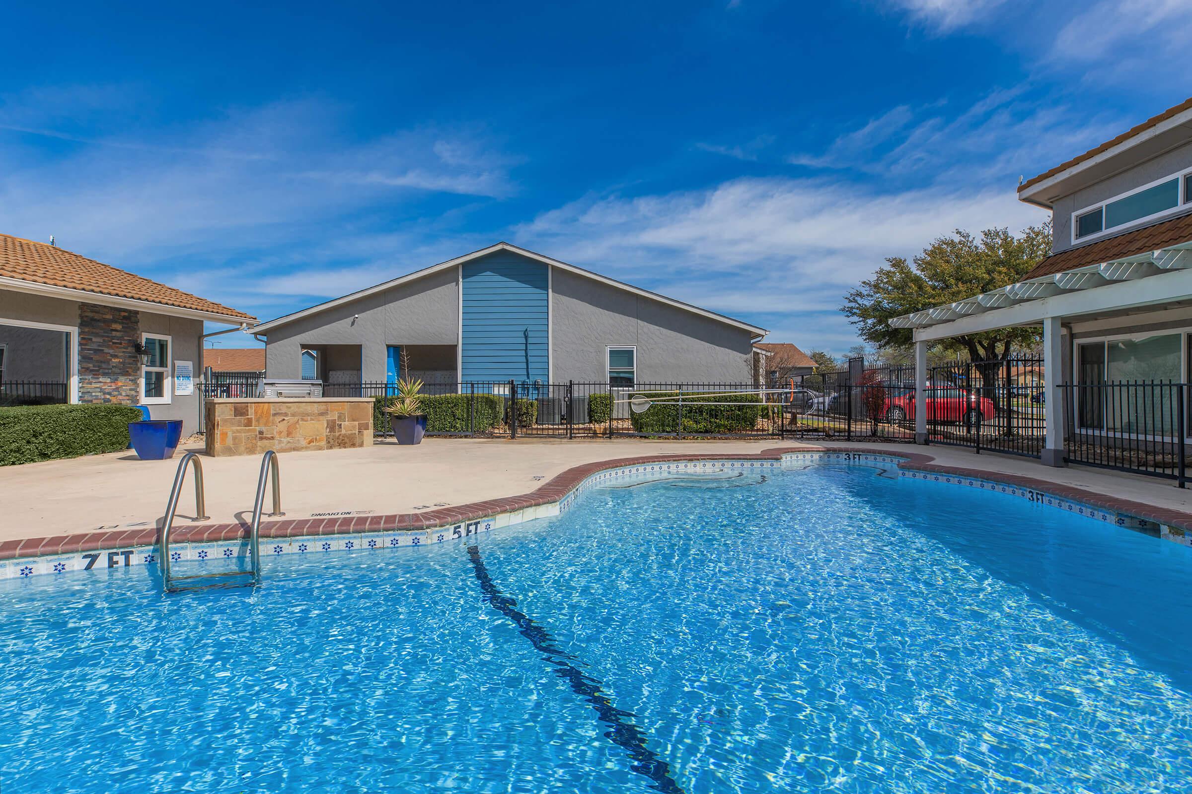 A clear blue swimming pool surrounded by a concrete deck. In the background, there are two buildings with a mix of stone and siding exteriors, along with green landscaping and tall trees. The sky is bright blue with a few clouds, creating a sunny atmosphere.