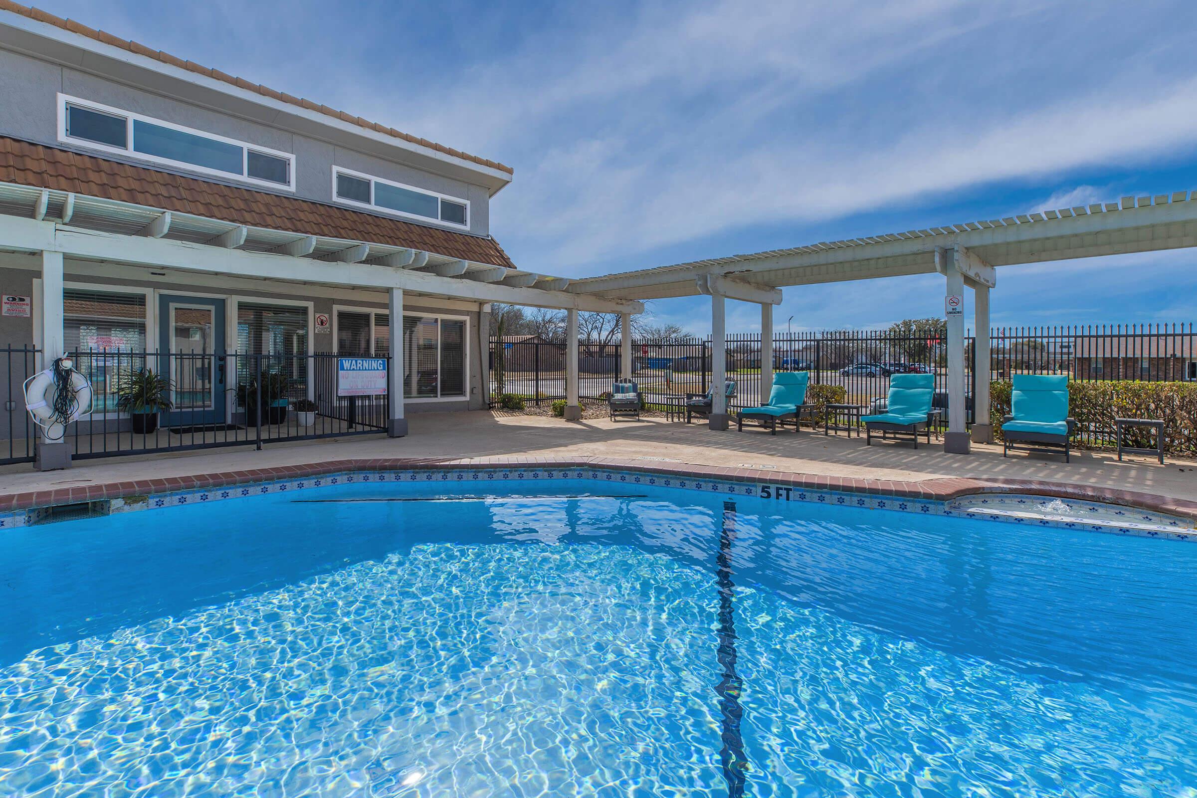 A clear blue swimming pool with a tiled edge, surrounded by lounge chairs under shaded pergolas. In the background, there's a modern building with large windows and a welcoming entrance. The sky is bright with a few clouds, creating a pleasant atmosphere for relaxation.