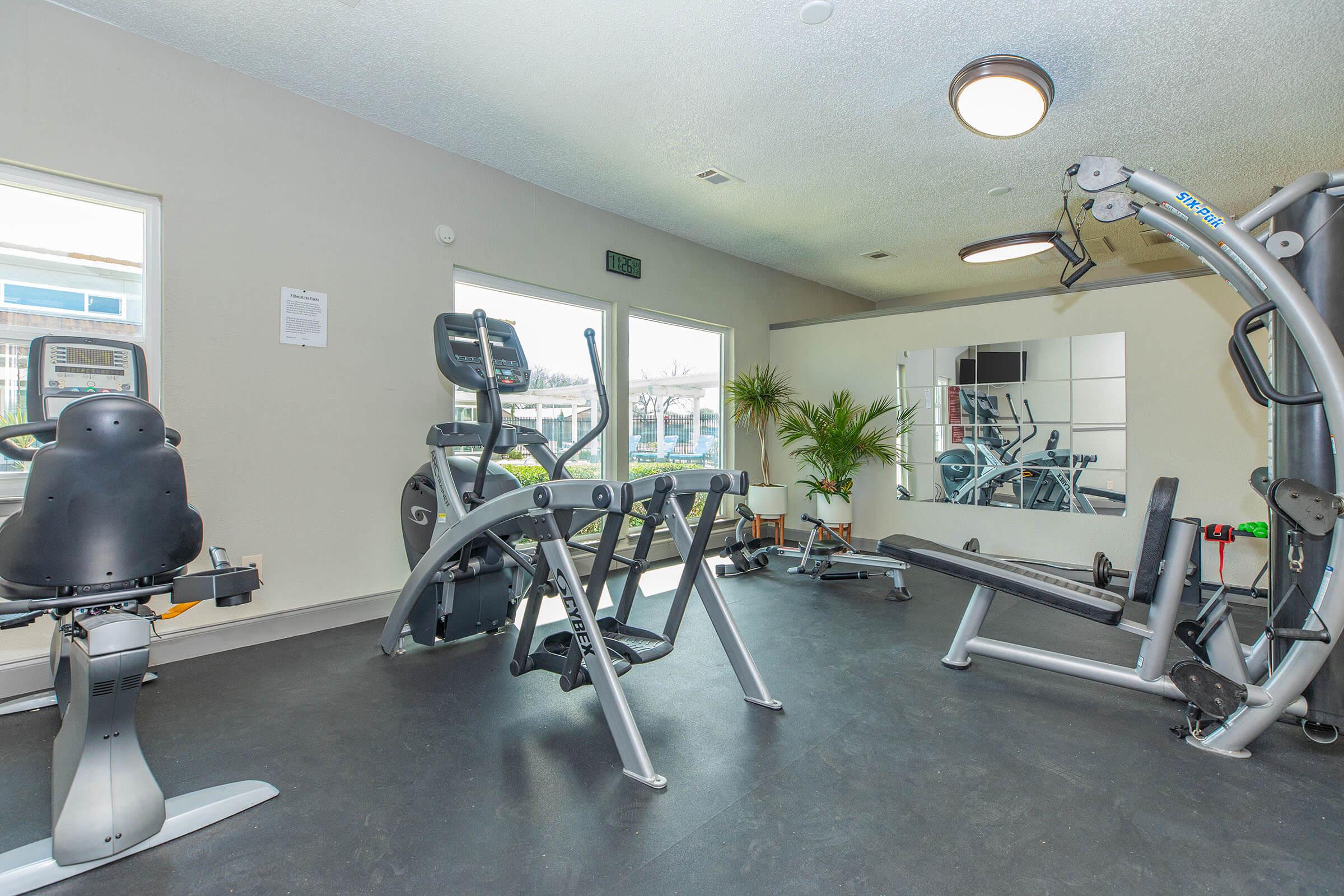 A modern gym interior featuring various exercise equipment, including a stationary bike, weight bench, and resistance machines. Large windows provide natural light, and a mirror is visible on one wall. The floor is dark, and there are plants for decoration, creating an inviting fitness space.