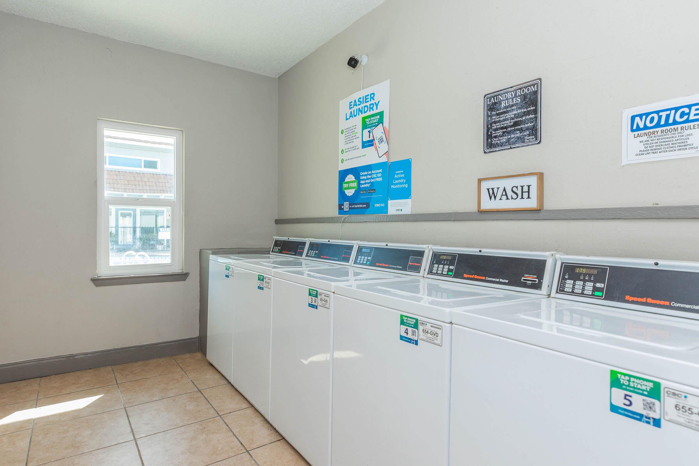 A clean, well-lit laundry room featuring multiple white washing machines in a row. The wall displays signs with laundry instructions and rules. A window allows natural light, and there is a sign labeled "WASH" on the wall. The floor is tiled, enhancing the space's neat and organized appearance.