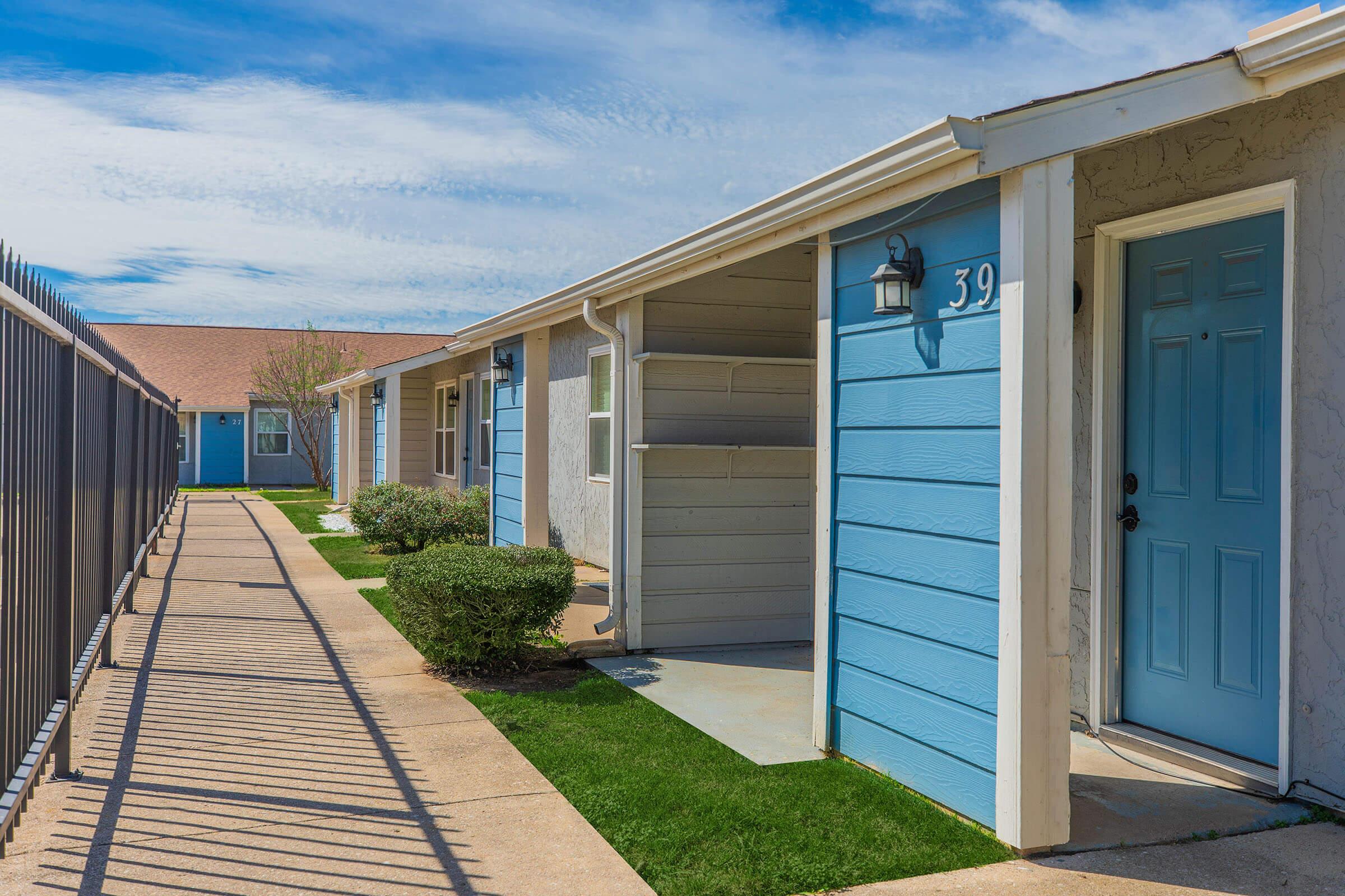 A row of residential buildings with light blue exteriors and cream-colored doors. Each unit features a small entry area with a pathway leading to the door. The scene includes manicured lawns and a clear blue sky, creating a serene atmosphere. A fence is visible in the foreground.