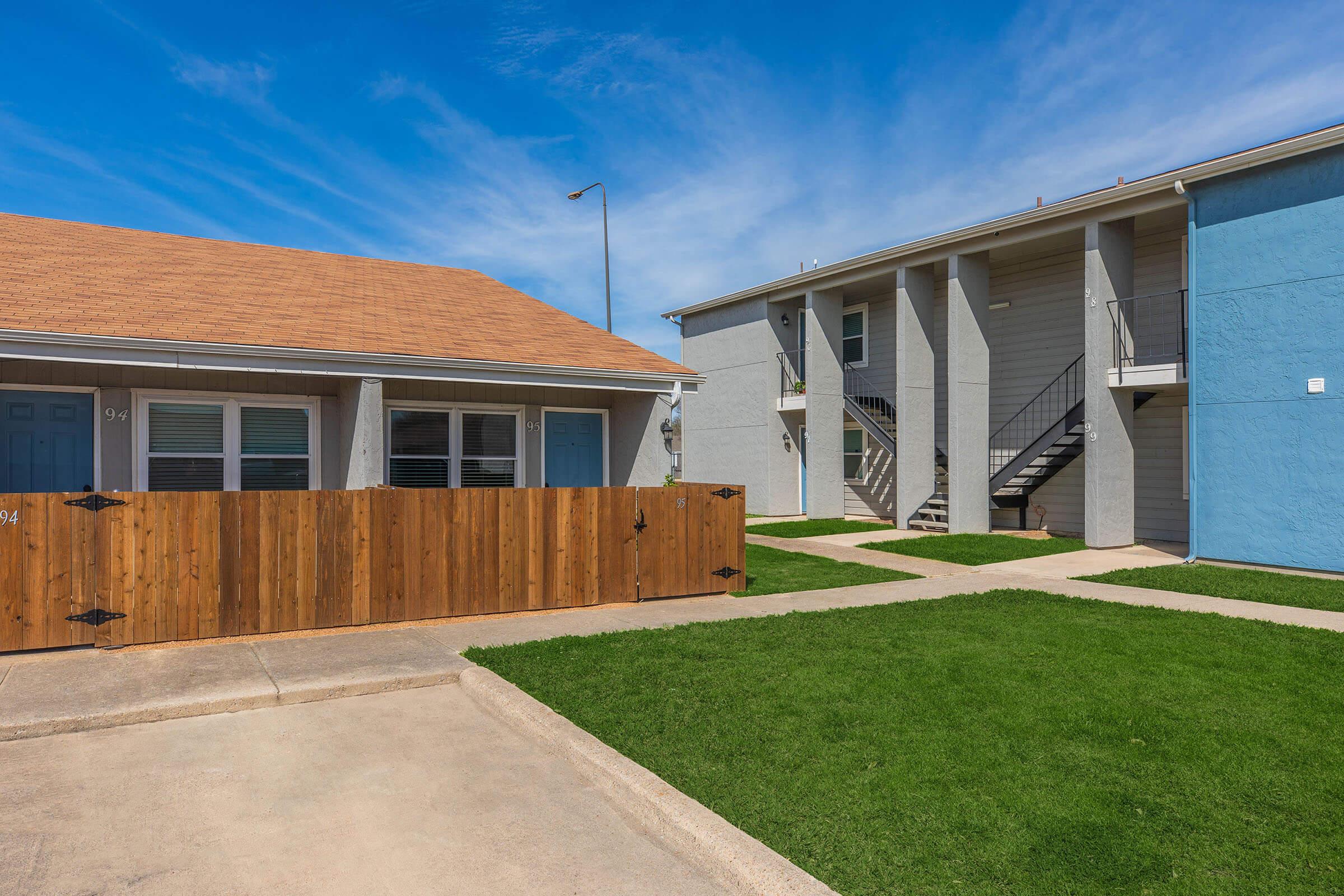 Two residential buildings side by side, one with a tan roof and a white exterior featuring a fenced yard, and the other a blue building with stairs leading to upper units. The scene shows well-maintained green lawns under a clear blue sky.