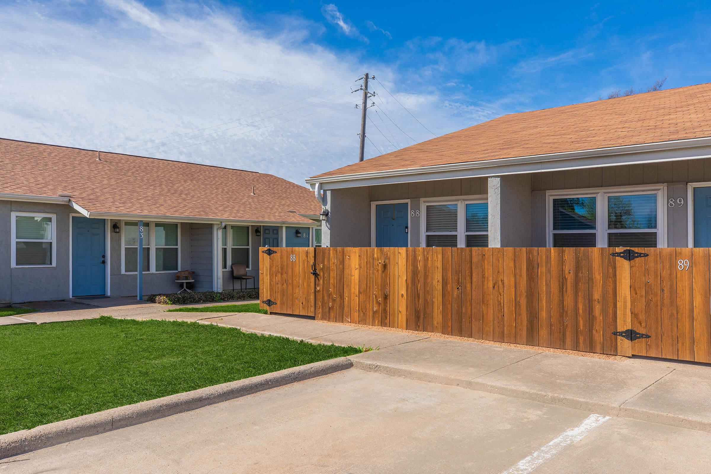 Two small, single-story buildings with light blue and gray exteriors, featuring sloped roofs. A wooden fence with metal accents separates the two units. The foreground shows a paved area with a grassy patch, and a clear blue sky in the background. A telephone pole is visible in the distance.