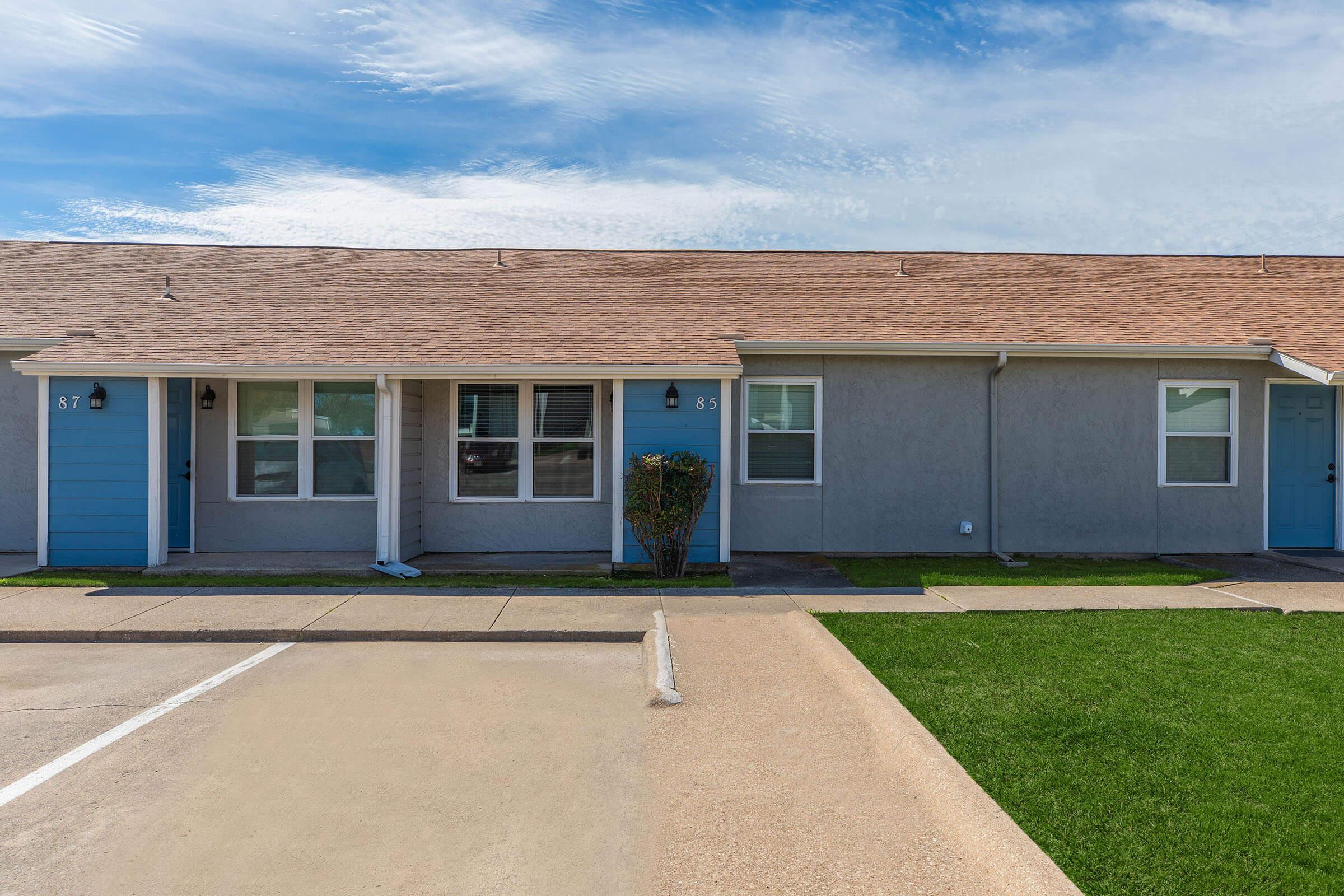 View of a row of single-story residential units with two doors visible, one light blue and the other gray. The sidewalk leads up to the doors, and there is a small shrub next to the blue door. The housing complex has a clean asphalt parking area and well-maintained green grass in front. The sky is clear with a few clouds.