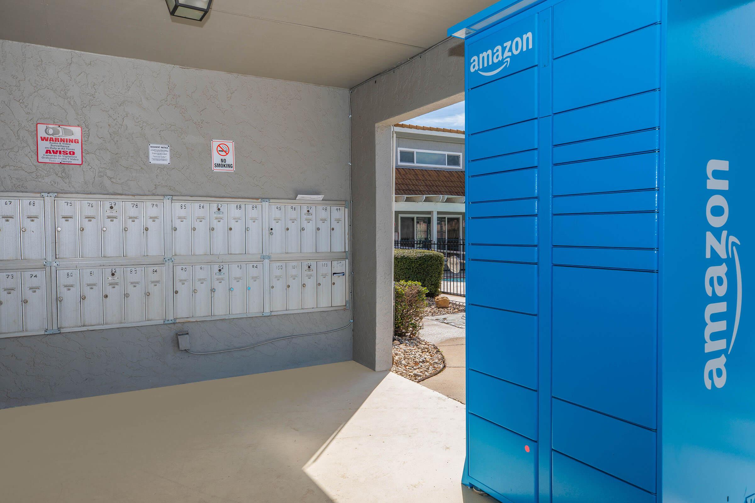 A brightly lit entrance with an Amazon locker on the right and a row of traditional mailboxes on the left. The scene is set in a well-maintained area, with landscaping visible outside. Signs are posted near the mailboxes, indicating regulations or warnings.