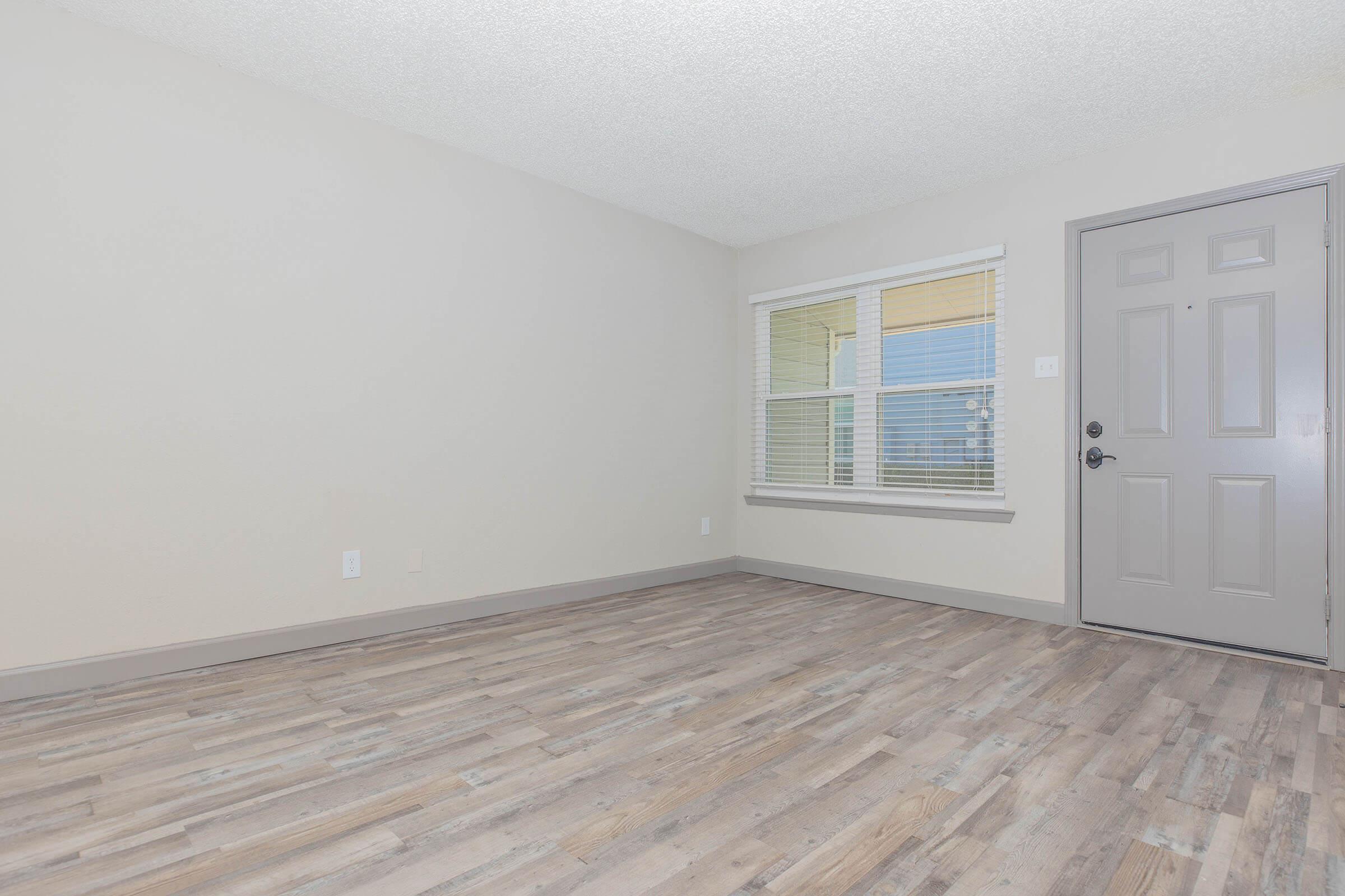 A spacious, empty room with light-colored walls and a textured ceiling. There is a window with closed blinds on the left side, allowing soft natural light. The floor features wood-like laminate, and a closed gray door is visible to the right. The room is clean and ready for furnishing.