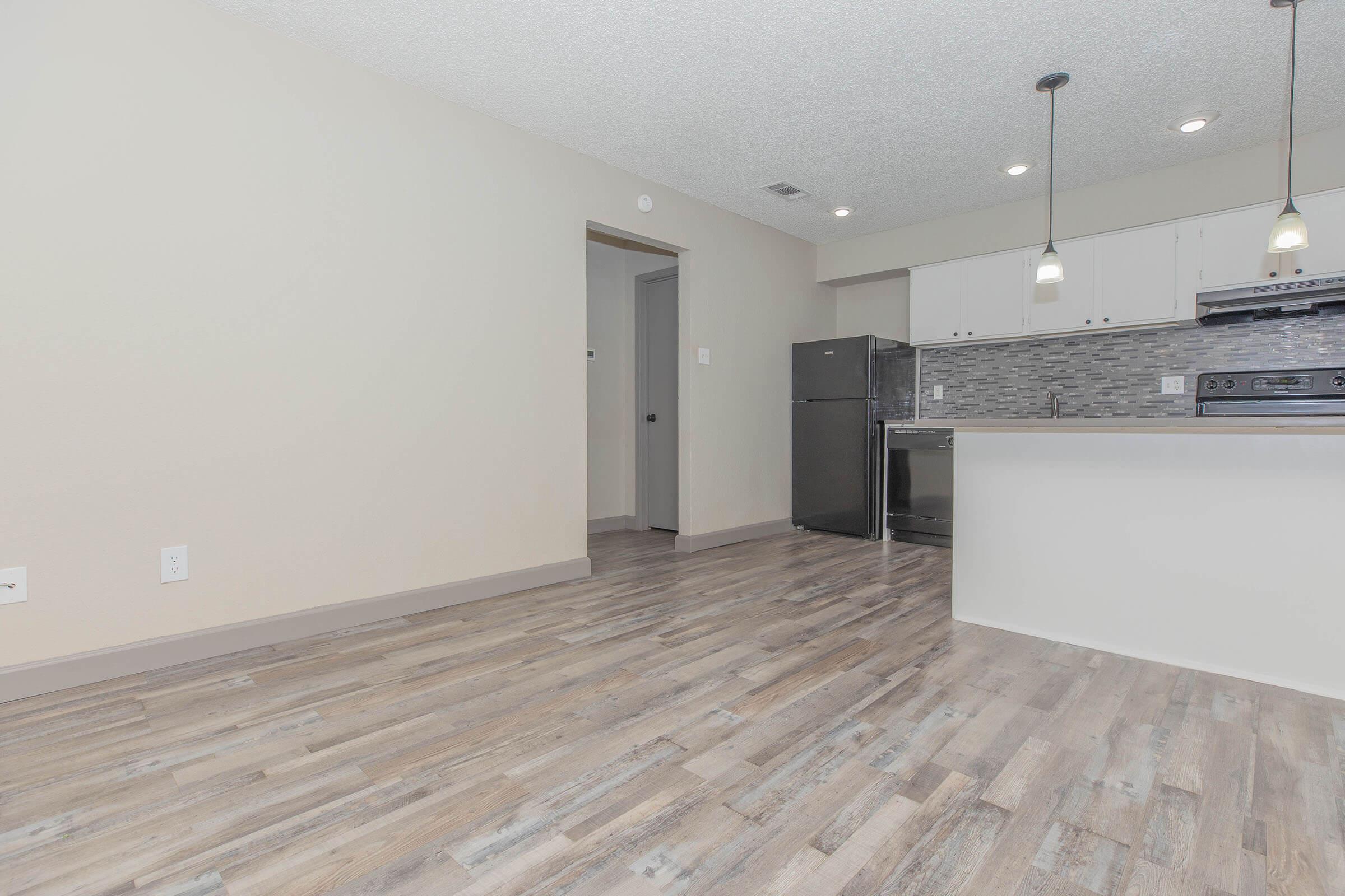 Interior of a modern apartment showcasing an open-layout kitchen and living area. The kitchen features a black refrigerator, dark cabinetry, and light-colored walls. Light wood-like flooring extends throughout the space, with pendant lighting hanging over the kitchen counter. The area is bright and inviting.