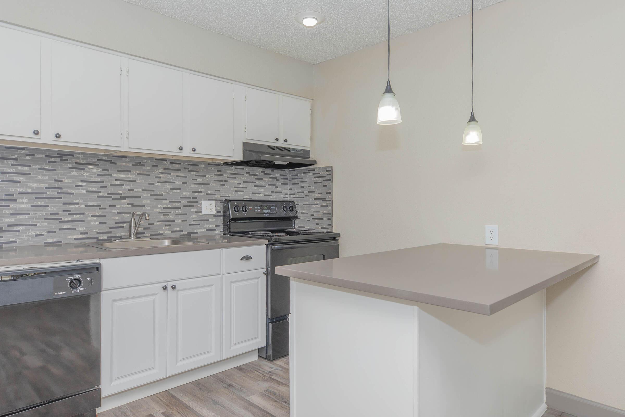 A modern kitchen featuring white cabinetry, stainless steel appliances, a tiled backsplash in shades of gray, and a beige countertop. There are two pendant lights hanging above a small island, and the flooring is wood-like laminate. The overall design is clean and contemporary.