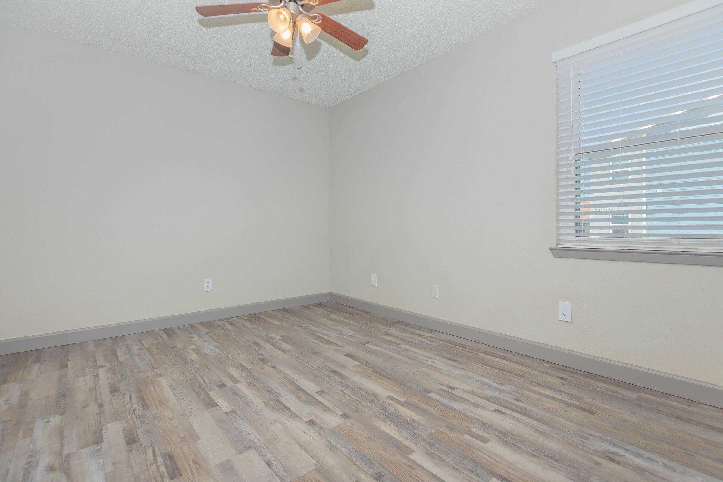 Empty room with light-colored walls and a ceiling fan with wooden blades. The floor features light brown laminate planks. A window with white blinds allows natural light to enter, and there are electrical outlets and light switches visible on the walls.