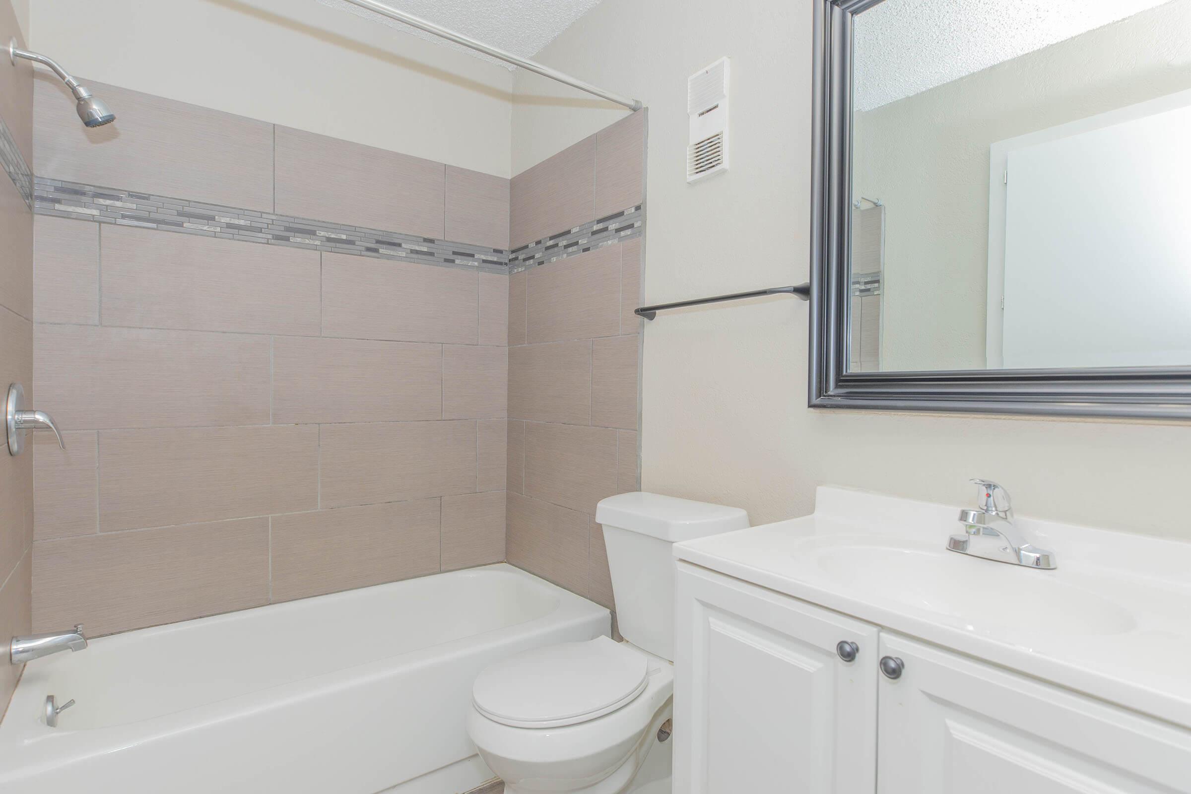 A clean, modern bathroom featuring a bathtub with a shower, beige tiled walls with a decorative strip, a white toilet, and a white vanity with a sink. A large mirror above the vanity reflects the space, and there is a light fixture on the wall. The overall color scheme is neutral and contemporary.