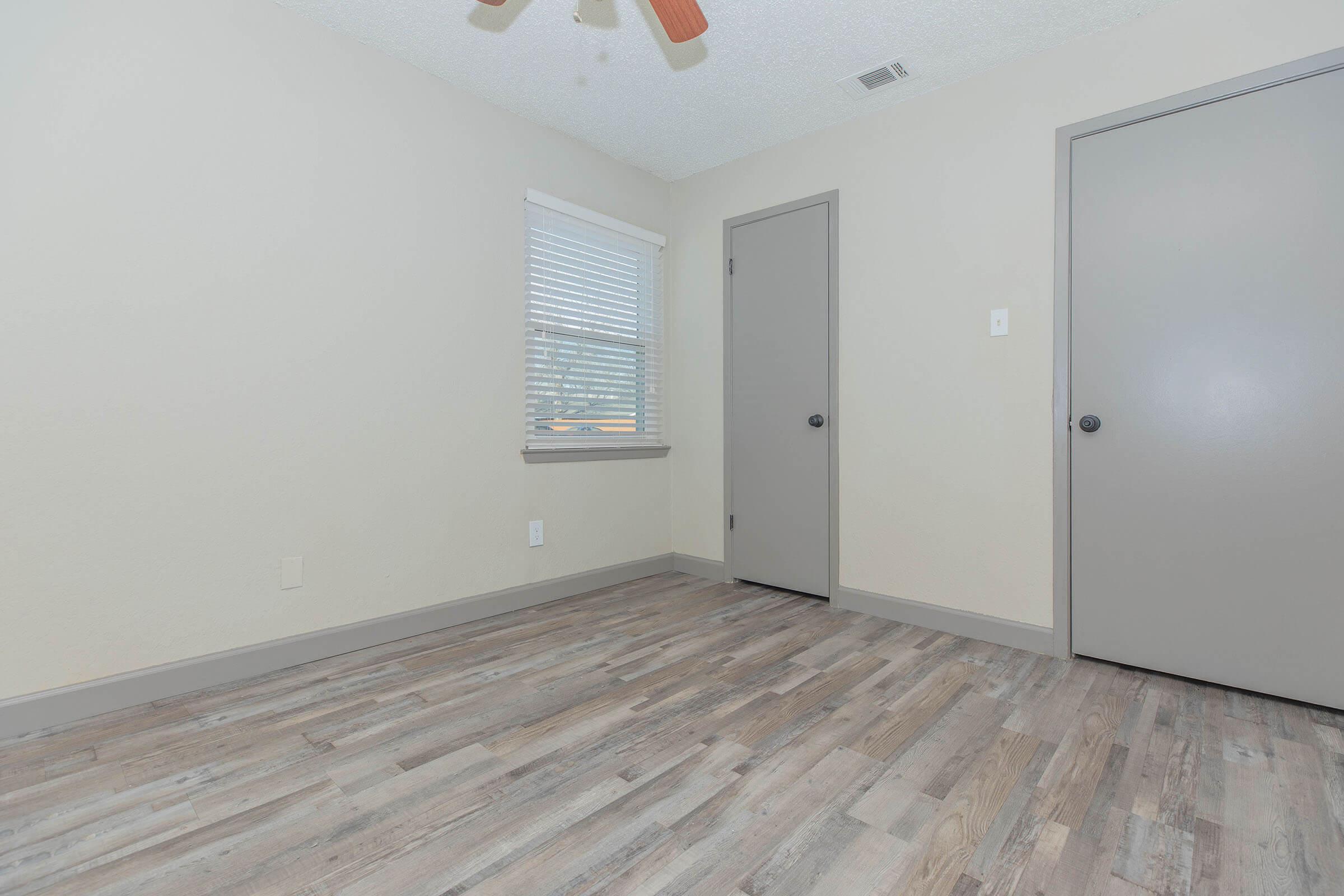 A small, empty room with light-colored walls and textured wooden flooring. There is one window with blinds, and two closed doors on opposite walls. The ceiling has a ceiling fan, and the room is well-lit by natural light, creating a bright and airy atmosphere.