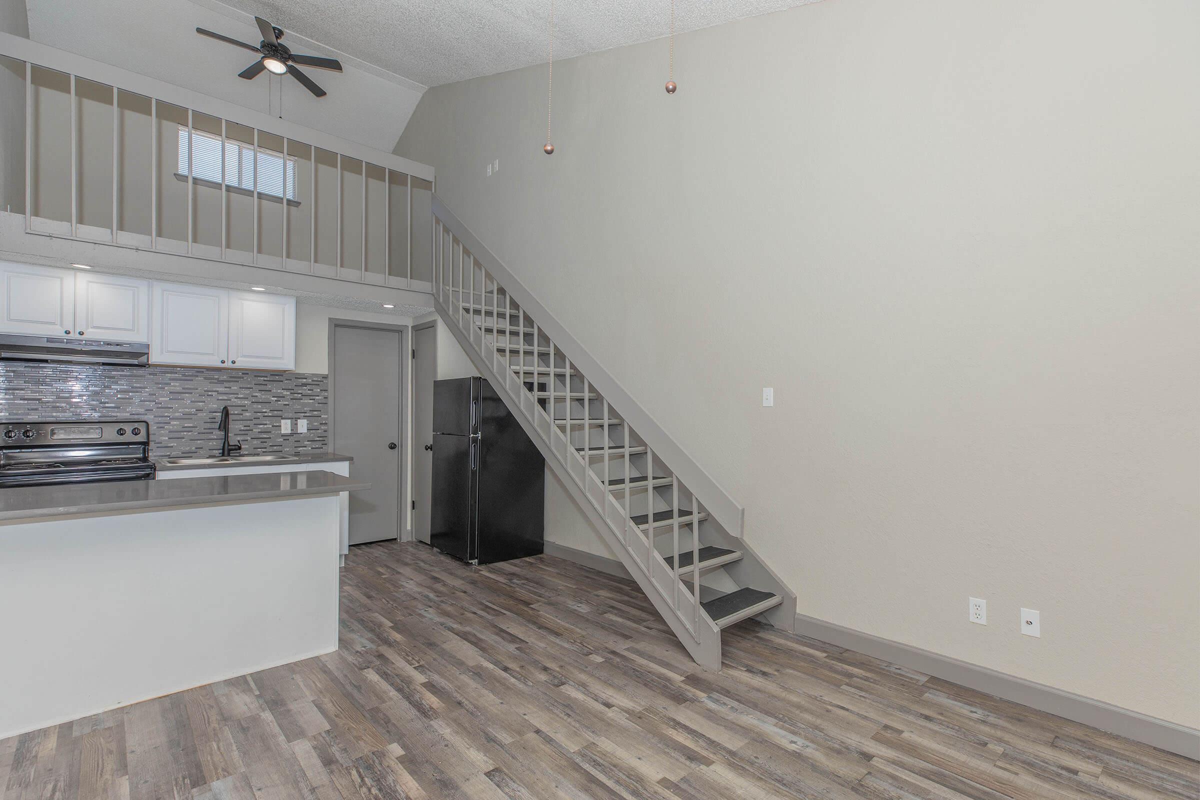 A modern, open-concept living space featuring a kitchen with white cabinets, a black fridge, and a stainless steel oven. A staircase leads to a loft area, and large windows provide natural light. The flooring is a light wood style, enhancing the contemporary aesthetic, with neutral-colored walls creating a welcoming atmosphere.