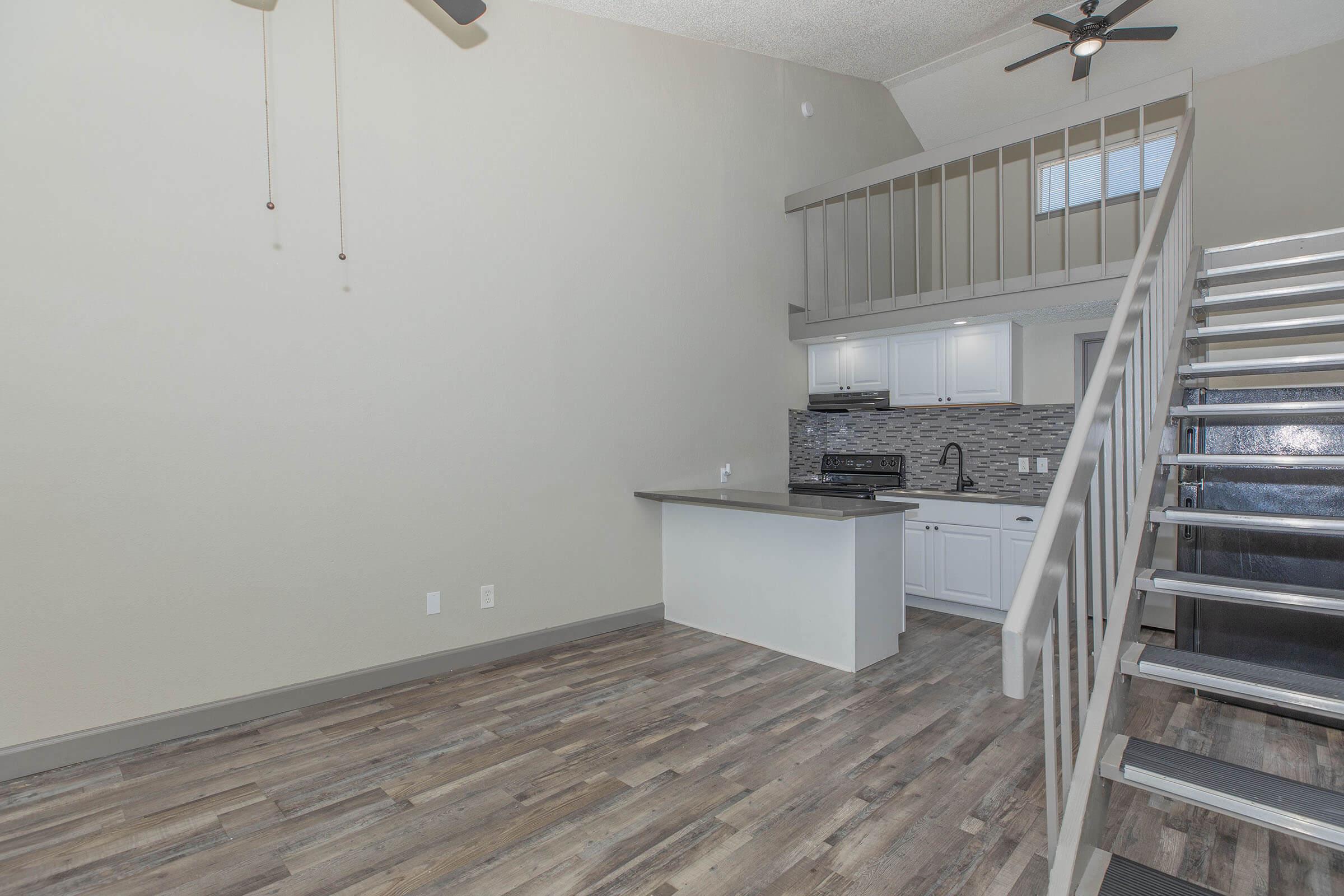 A spacious indoor area featuring a modern kitchen with white cabinetry, a dark countertop, and a tiled backsplash. The open layout includes a staircase leading to a loft or upper level, and large windows allow natural light to fill the space. The flooring is hardwood-style, adding warmth to the room.