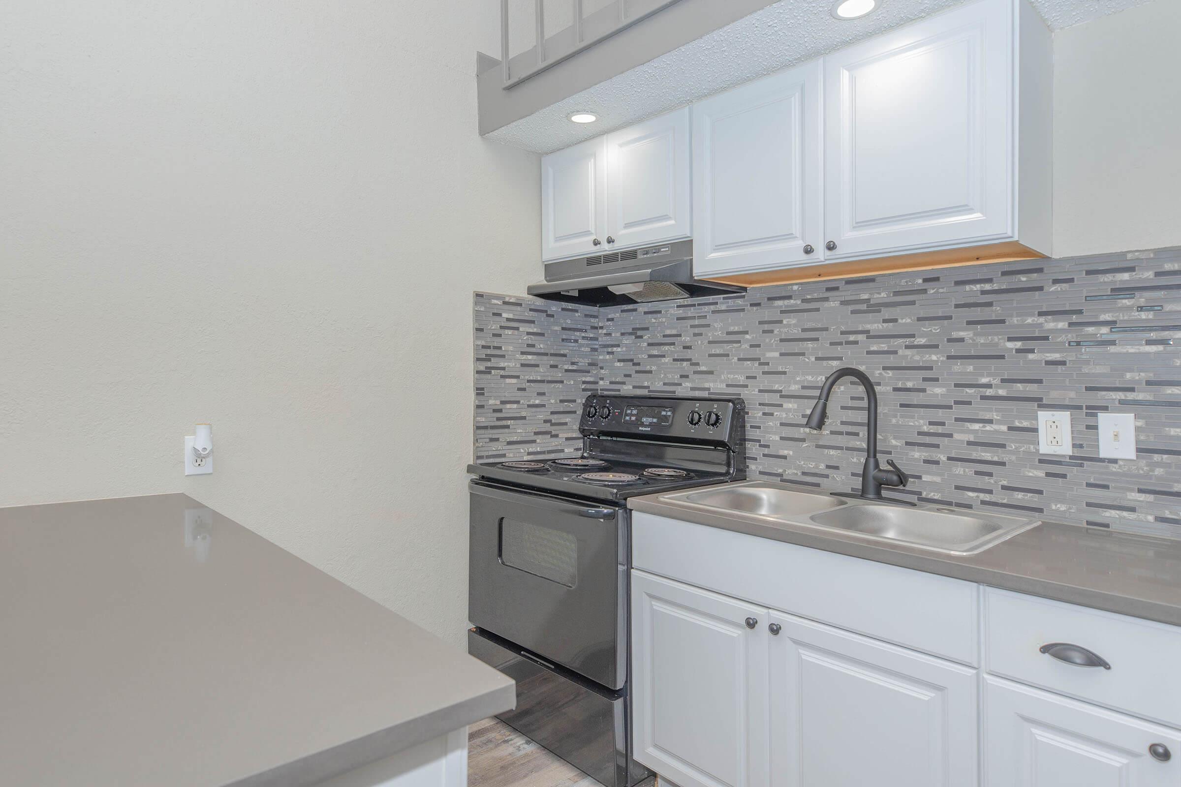 A modern kitchen featuring white cabinetry, a sleek gray countertop, stainless steel appliances, and a decorative backsplash with gray and black tiles. The image shows an oven, a double sink, and an overhead range hood in a well-lit space. The overall design is clean and contemporary.