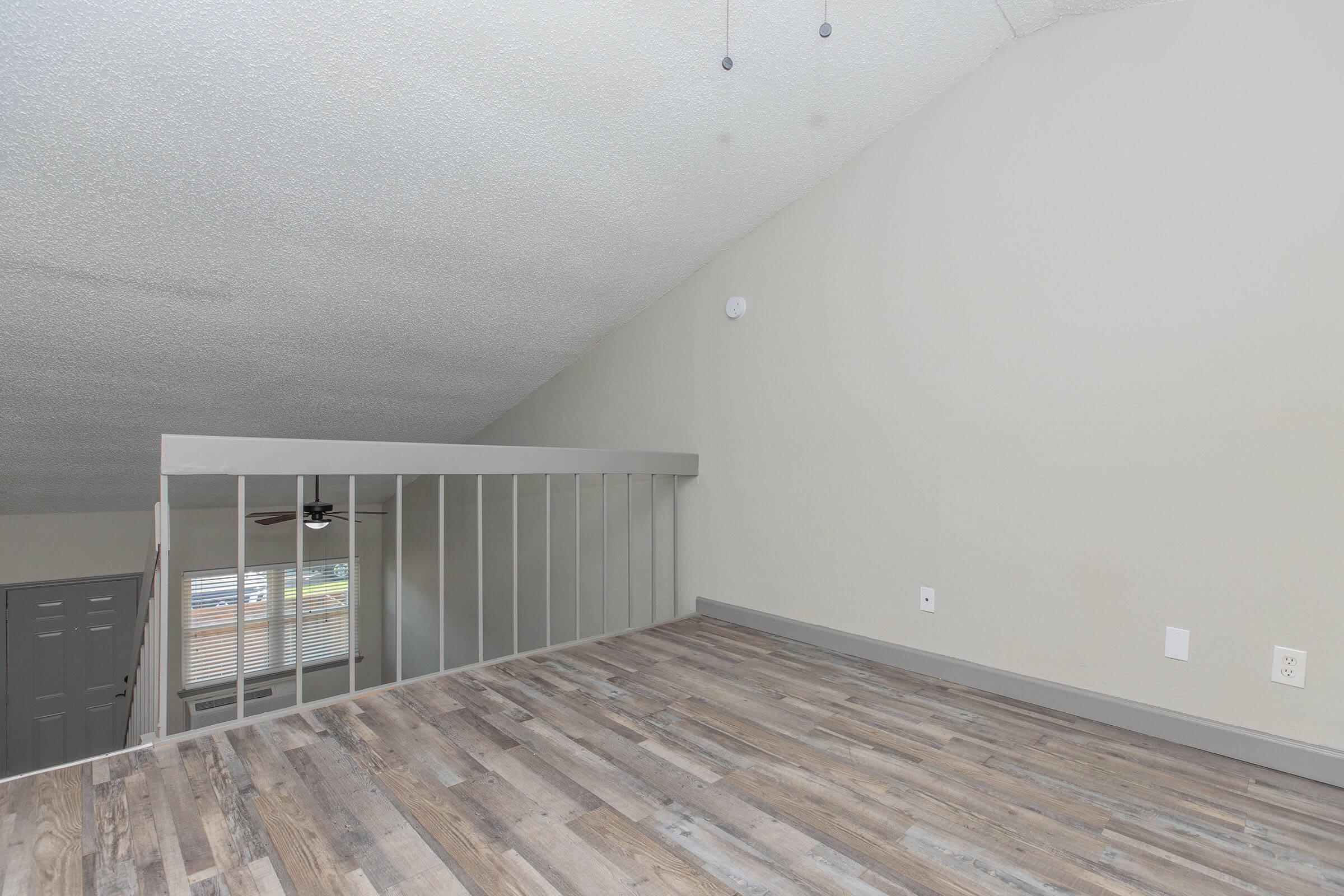 Interior view of a spacious room with a high ceiling, featuring light-colored walls and a wooden floor. A loft area is visible with a railing, and a ceiling fan is mounted above. Large window allows natural light to enter, creating an airy atmosphere.