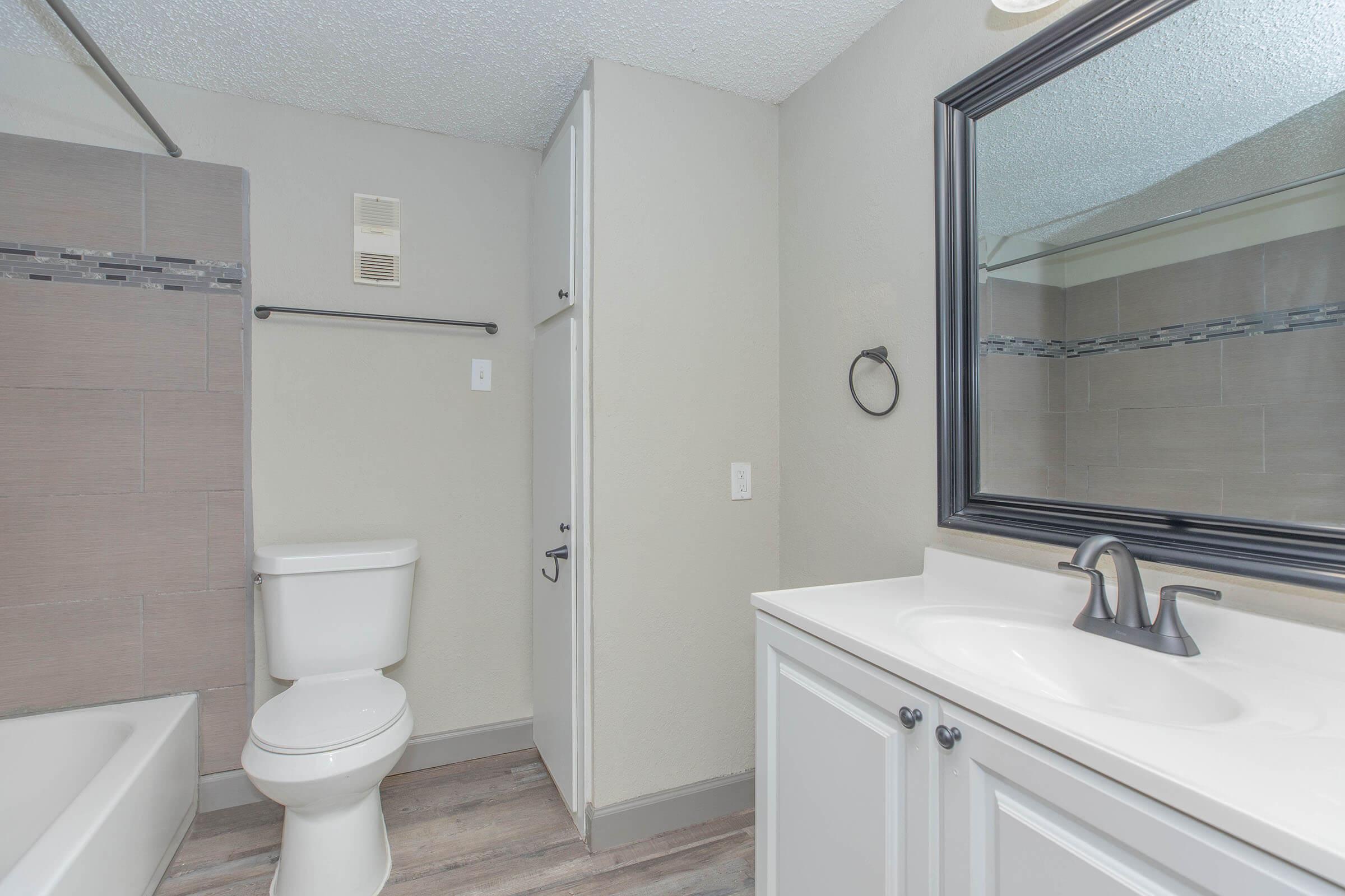 A modern bathroom featuring a white vanity with a sink, a large mirror, a toilet, and a bathtub with a gray tile surround. The walls are painted light gray, and there is a towel rack and ventilation grill visible. The flooring is a light wood look laminate.