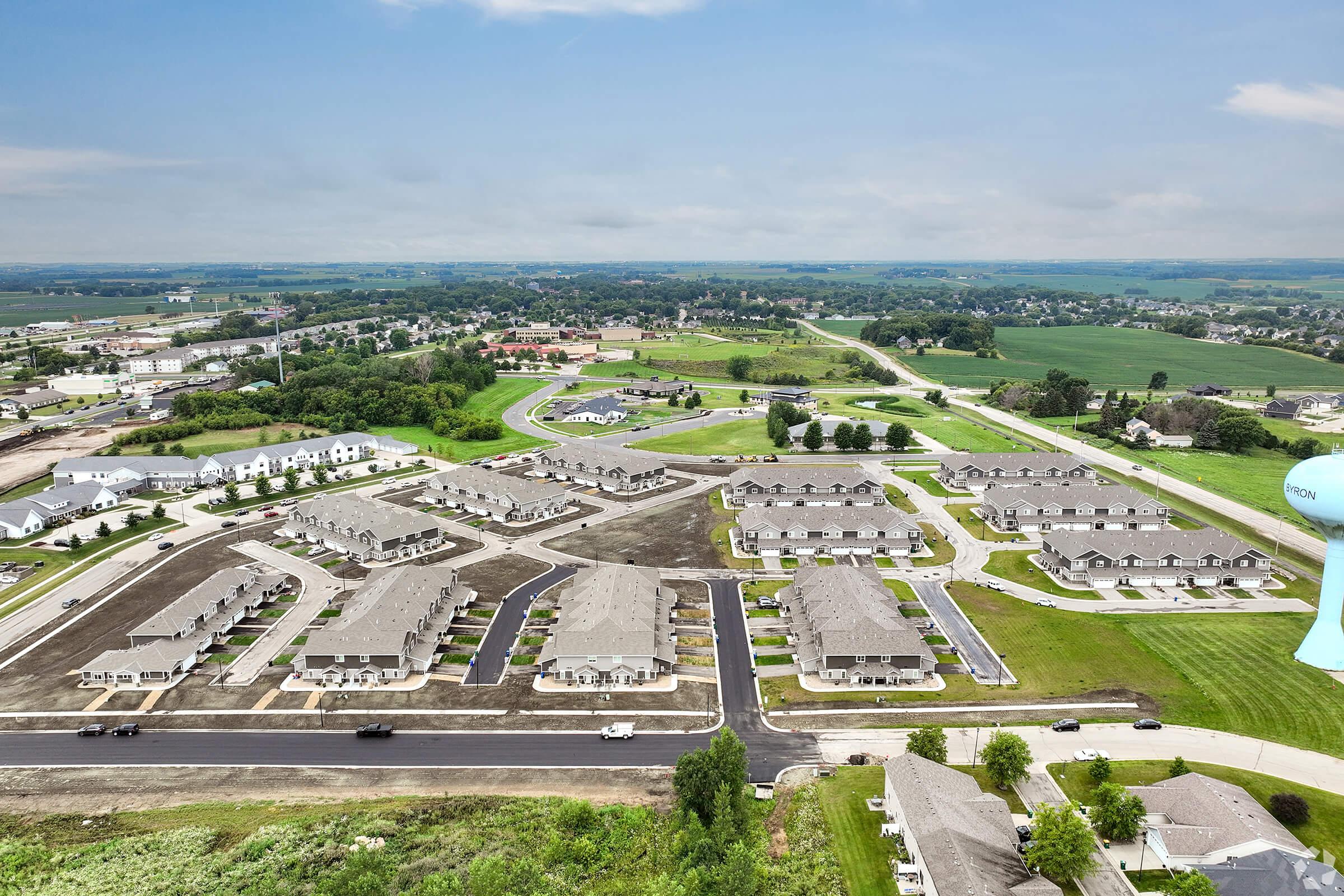 Aerial view of a residential area featuring multiple buildings arranged in neat rows, surrounded by green fields and roads. The landscape includes undeveloped land, a water tower in the distance, and houses within a suburban setting. The sky is partly cloudy, suggesting a pleasant day.