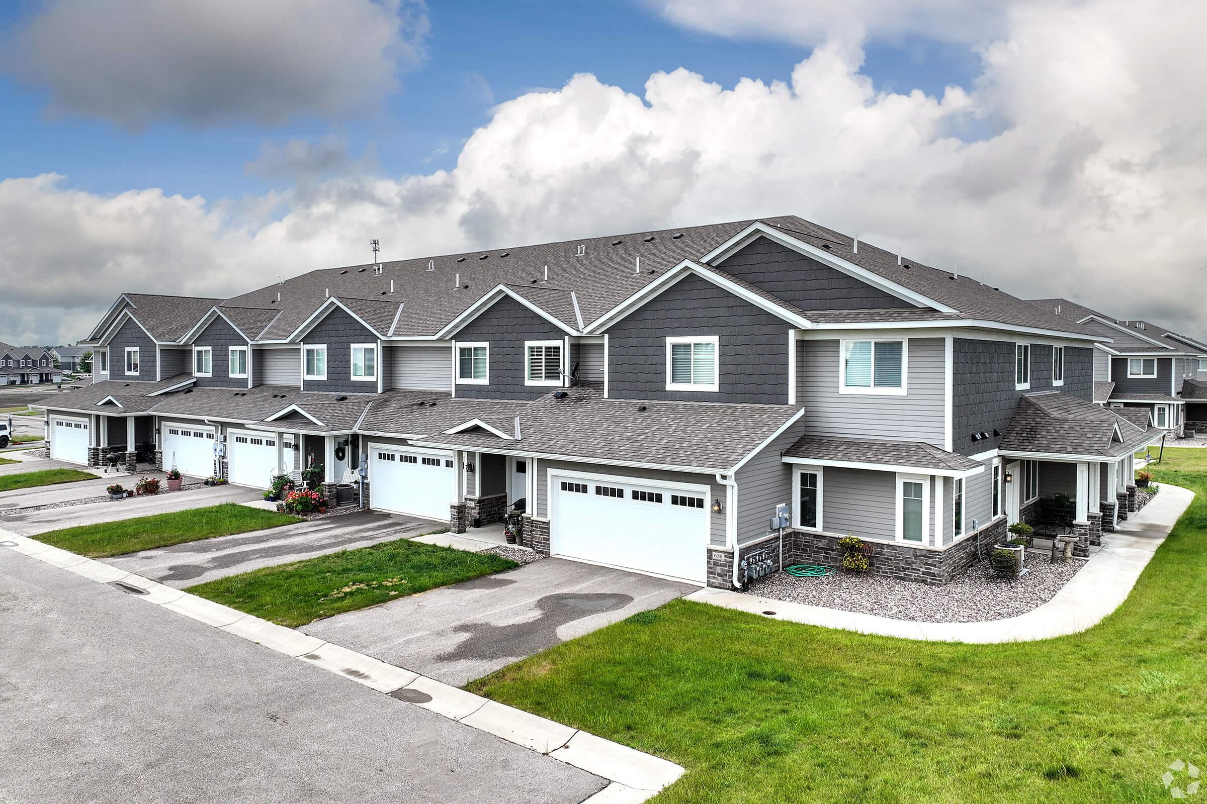 Row of modern townhouses with gray exteriors and garages, set against a cloudy sky. The grassy area in front features neatly trimmed lawns and flower pots. The pavement is visible, leading to the driveways, creating a tidy suburban atmosphere.