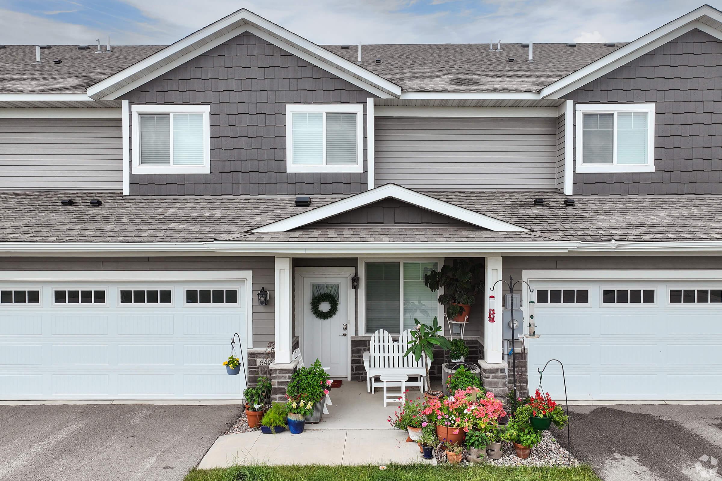 A well-maintained townhouse exterior featuring a gray façade with white trim. The entrance includes a decorative wreath on the door and a cozy white bench surrounded by vibrant potted plants and flowers. The garages are visible, and the overall scene is inviting and neat.