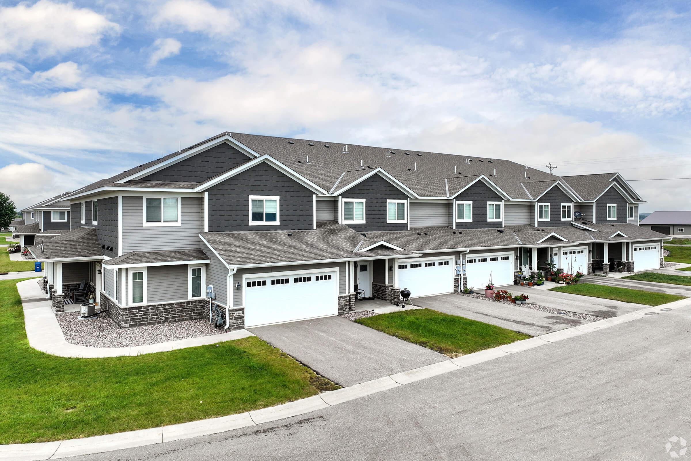 A row of modern, multi-story townhouses with gray exteriors and garages, situated on a well-maintained street. The lawns are neatly trimmed, and there are flower pots near the entrances, under a cloudy blue sky.