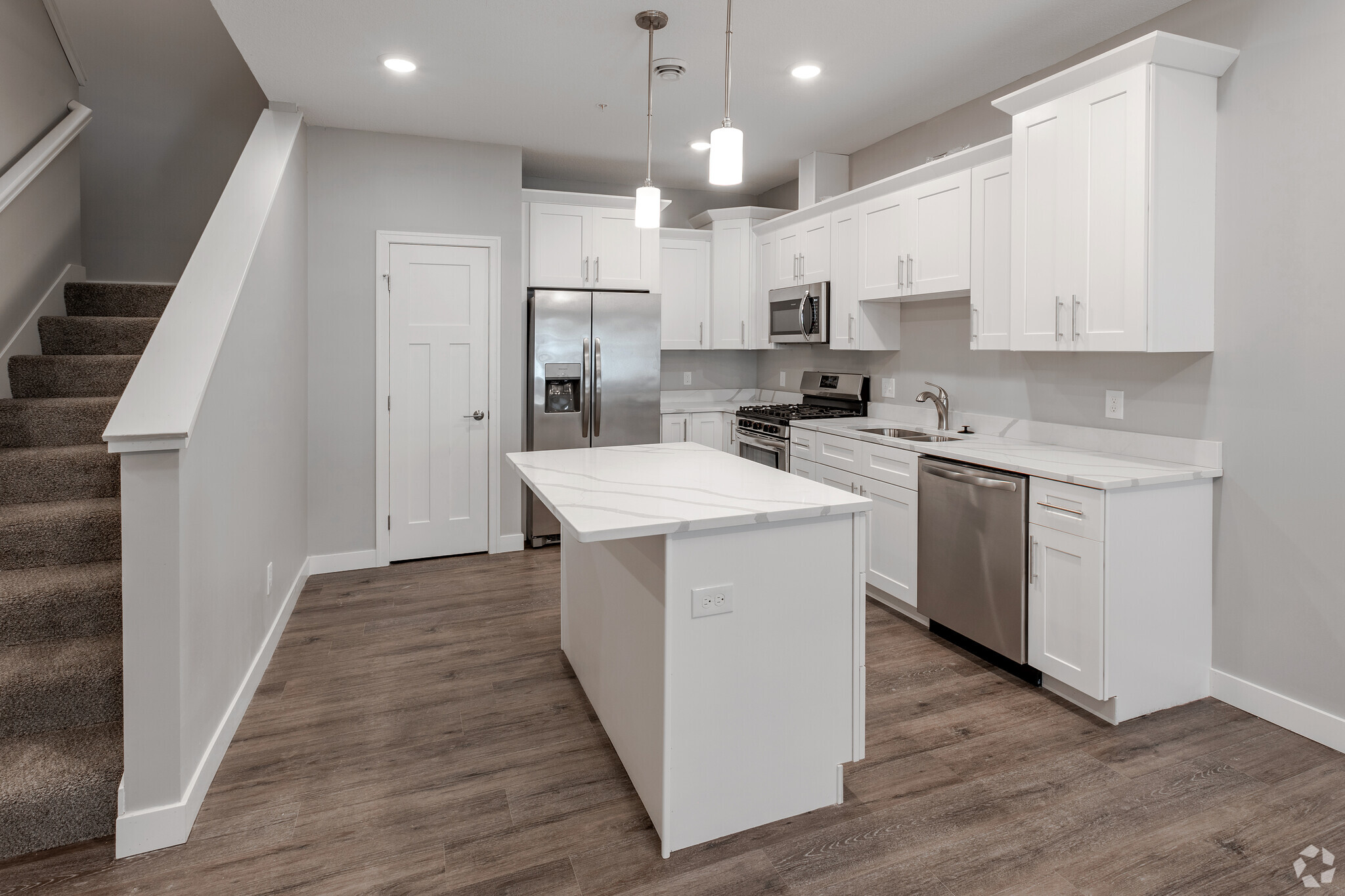 Modern kitchen interior featuring white cabinets, stainless steel appliances, and a large island with a marble countertop. The space has light wooden flooring and a staircase visible in the background. Bright overhead lights illuminate the area, creating a clean and inviting atmosphere.