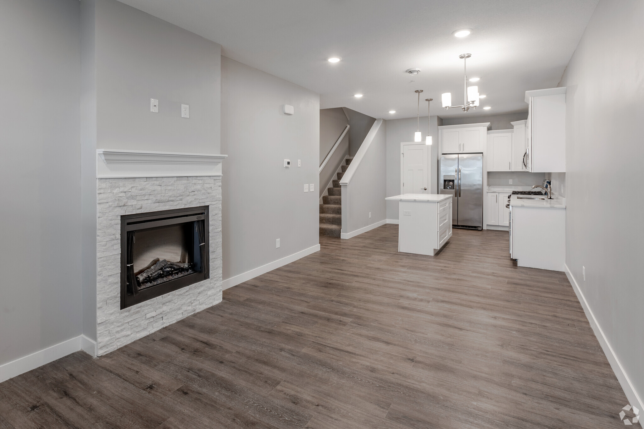 Modern open-concept living space featuring a corner fireplace, wood flooring, and ample natural light. The kitchen includes white cabinets and stainless steel appliances. A staircase leads to the upper level, and there's a neutral color palette throughout the room, creating a bright and inviting atmosphere.