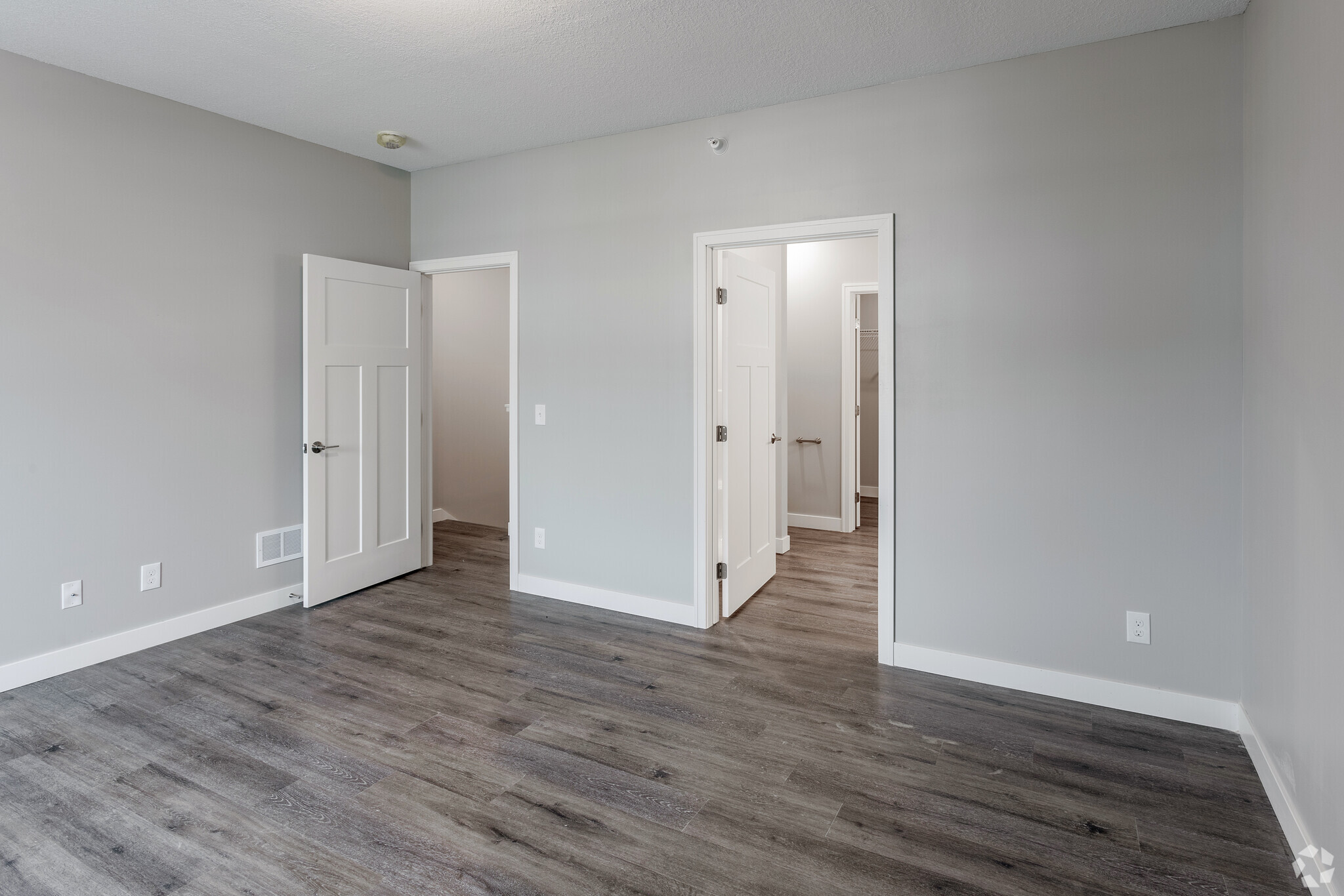 A spacious, modern room with light gray walls and hardwood-style flooring. It features three doors: one leading to a closet, another to a bathroom, and one open door leading to an adjacent room. Natural light illuminates the area, creating a bright and airy atmosphere.