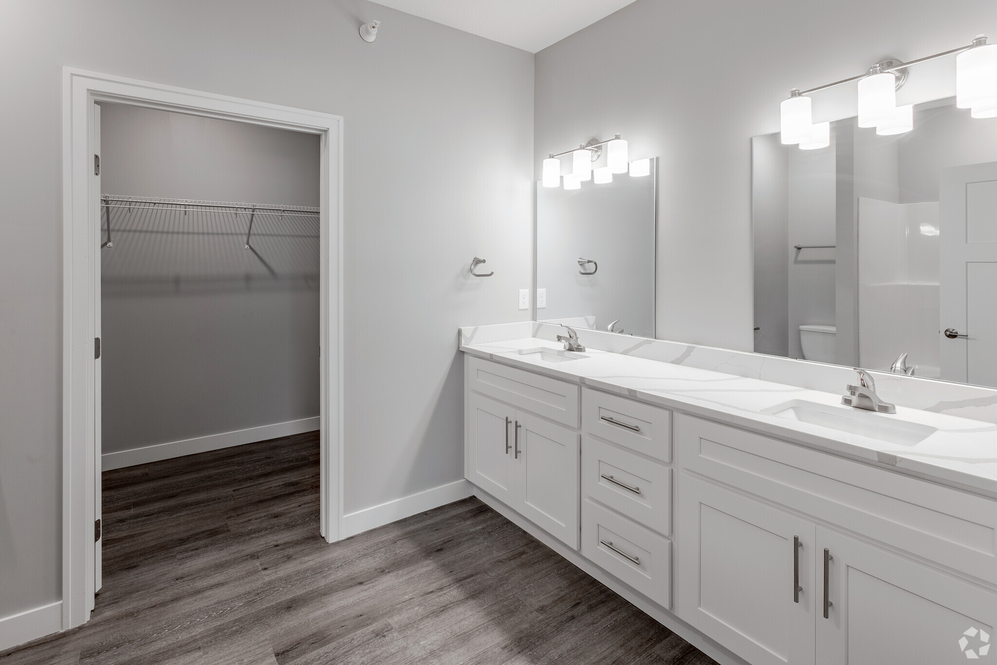 A modern bathroom featuring a double vanity with two sinks, stylish light fixtures above the mirrors, light gray walls, and wood-like flooring. To the left, there is a doorway leading to a spacious closet with a wire shelving unit.
