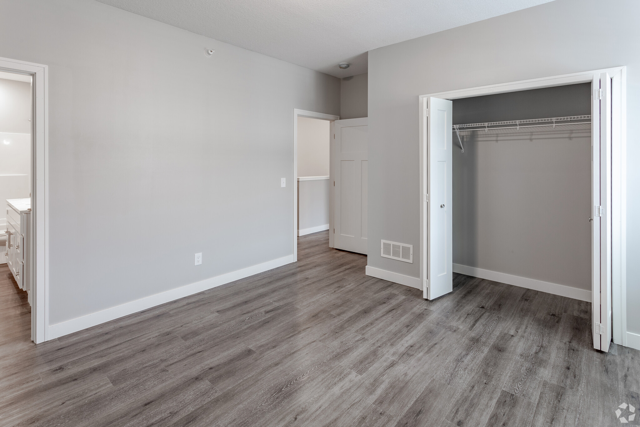 A spacious room with light gray walls and laminate flooring. Two open white doors lead to a closet and another room, suggesting a modern interior design. Natural light brightens the space, creating a fresh and inviting atmosphere.