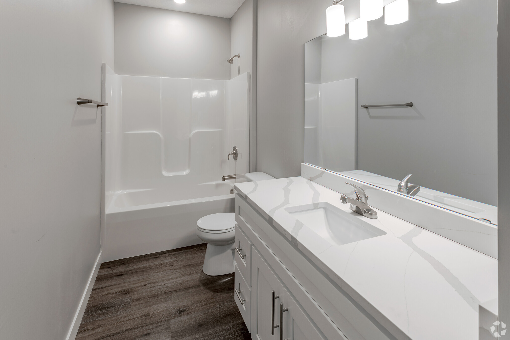 A modern bathroom featuring a white bathtub and shower combination, a sleek vanity with a white countertop, and a large mirror. The walls are painted in a light gray color, and the floor is covered with dark wood-like laminate. Bright overhead lighting illuminates the space.