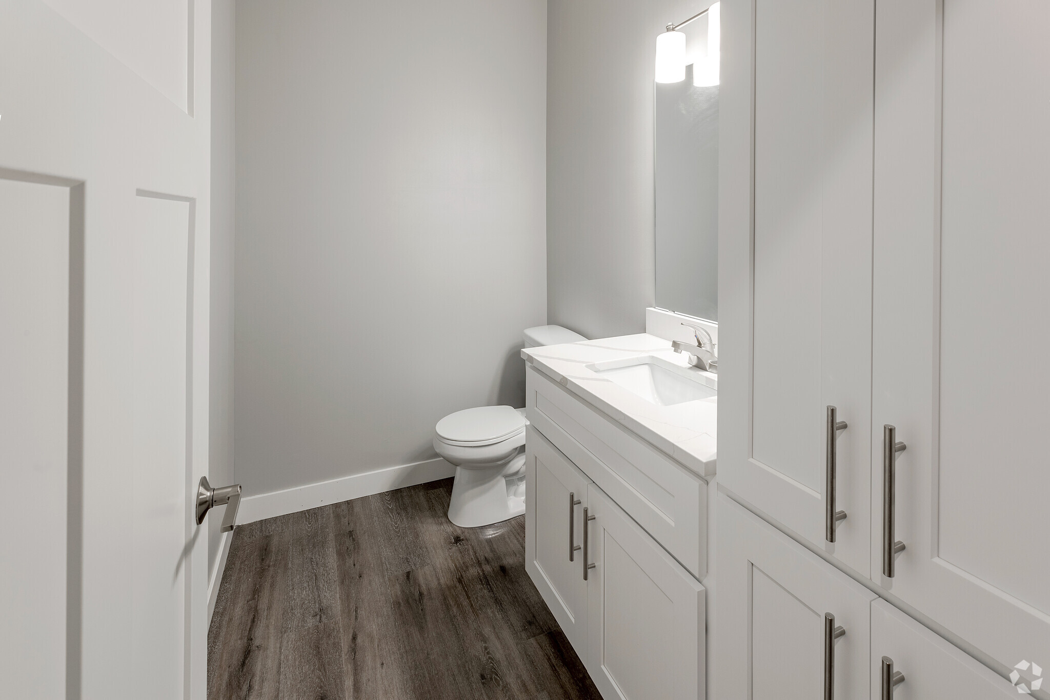 A modern bathroom featuring a white vanity with a marble countertop, a large mirror above it, and a toilet. The walls are painted in a light gray color, and the floor has dark wood-like planks. The space is well-lit with a ceiling light.