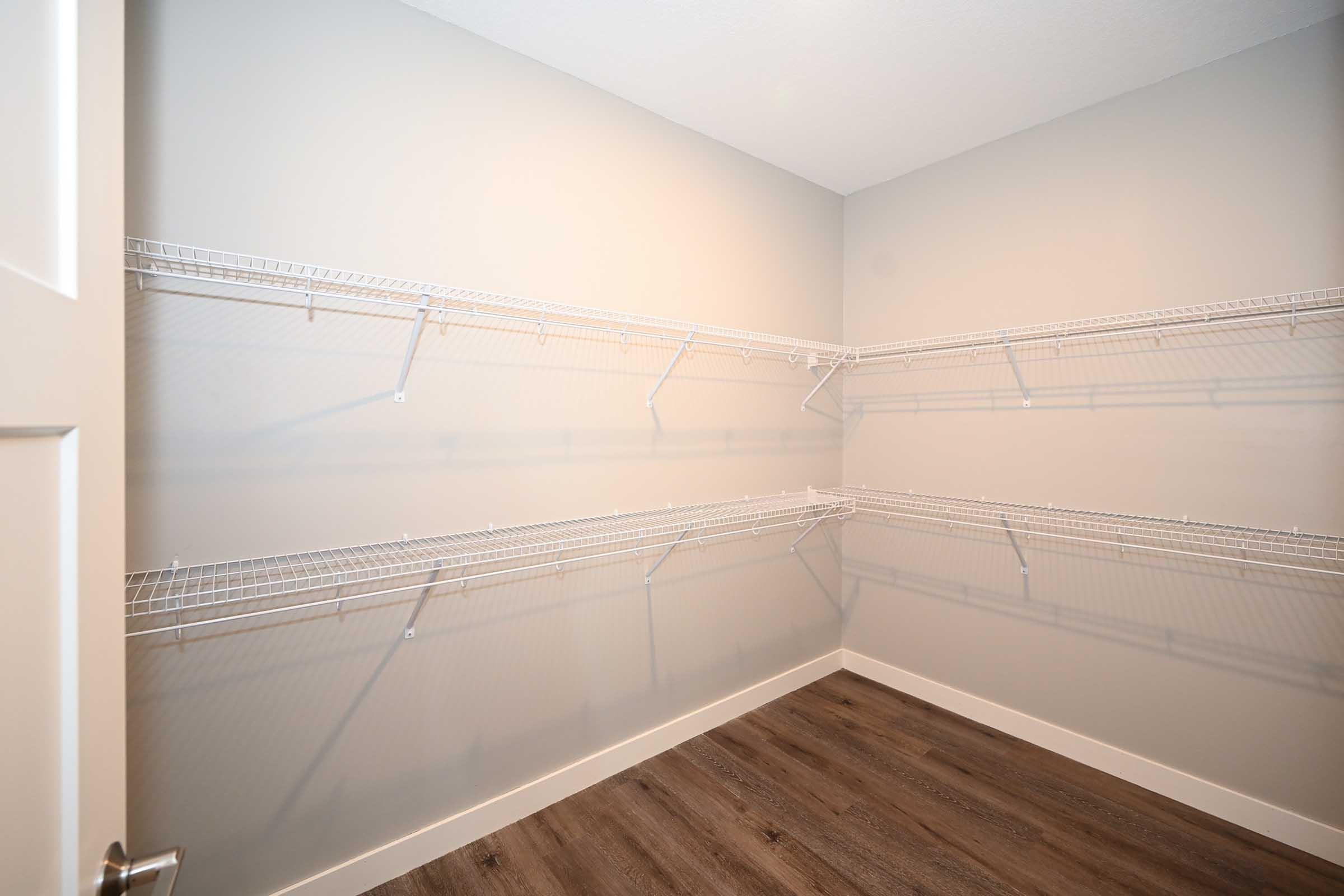 Empty closet with light gray walls and wooden flooring. Wire shelving units are mounted on the walls, providing storage space. The room is well-lit, highlighting the minimalistic design and clean lines. The door is partially open, showing the entrance to the closet.