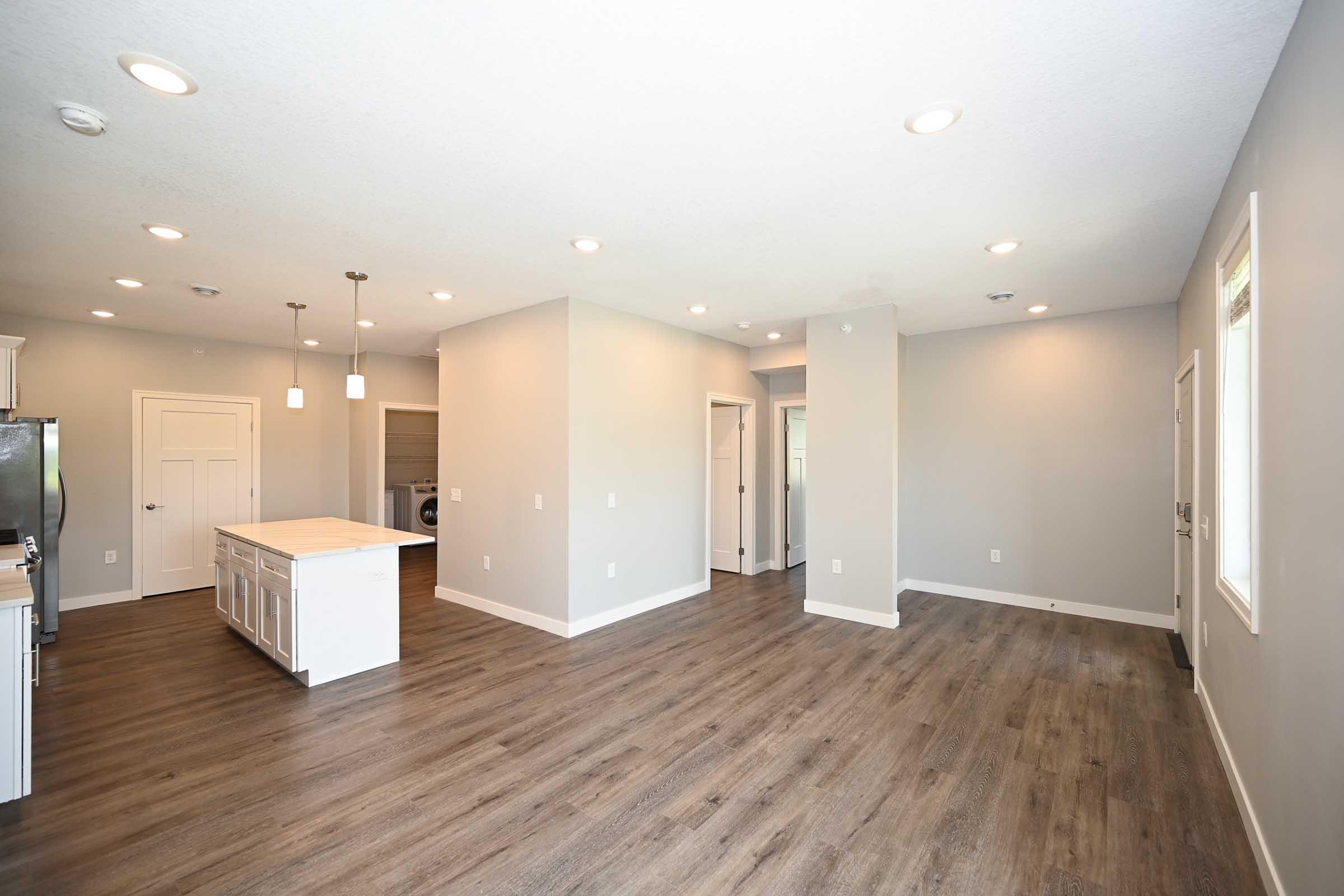 Spacious, modern interior of a home featuring an open layout with a kitchen island, gray walls, and wood-like flooring. Bright lighting illuminates the area. Doors lead to a laundry room and an exterior entrance, enhancing the functional design.
