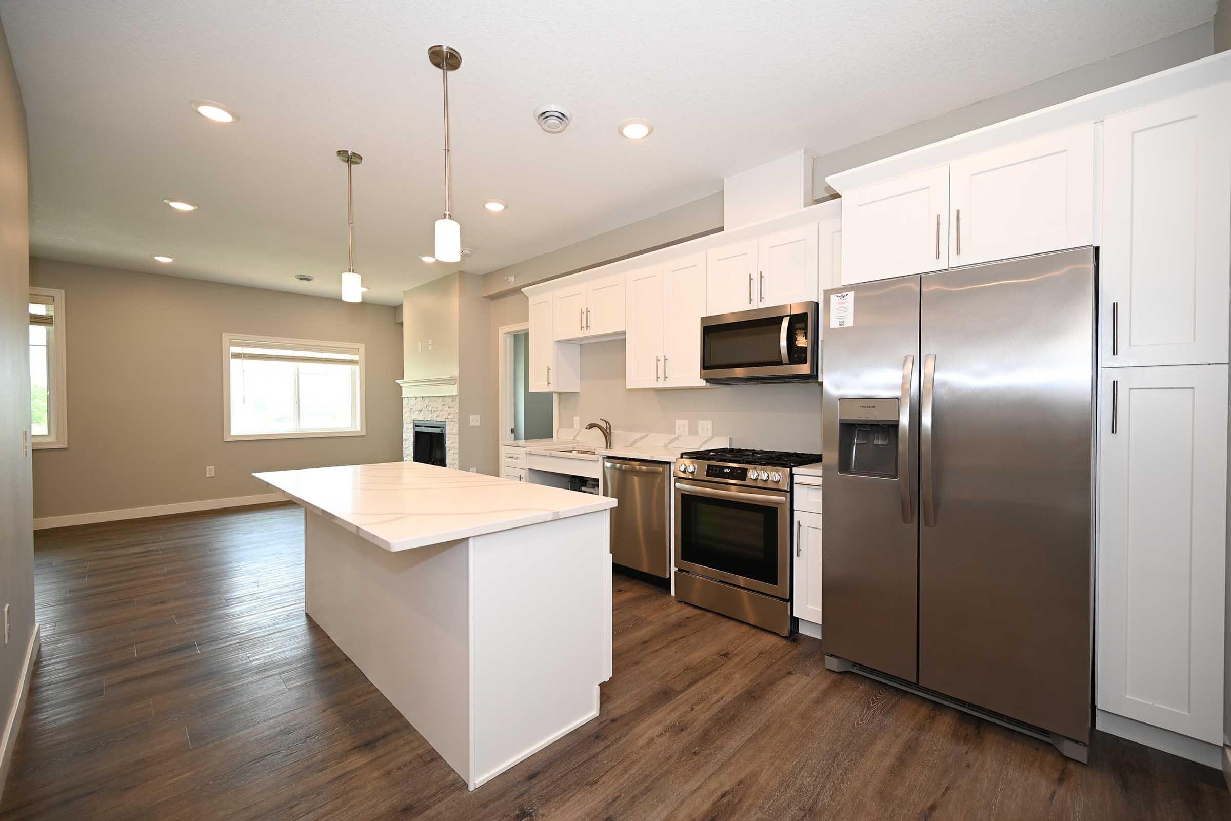 Modern kitchen with white cabinets, stainless steel appliances, and an island. Features include a gas range, microwave, and refrigerator, all set against a backdrop of laminate wood flooring and neutral-colored walls. The space is well-lit with recessed lighting and a window, creating a bright, open atmosphere.