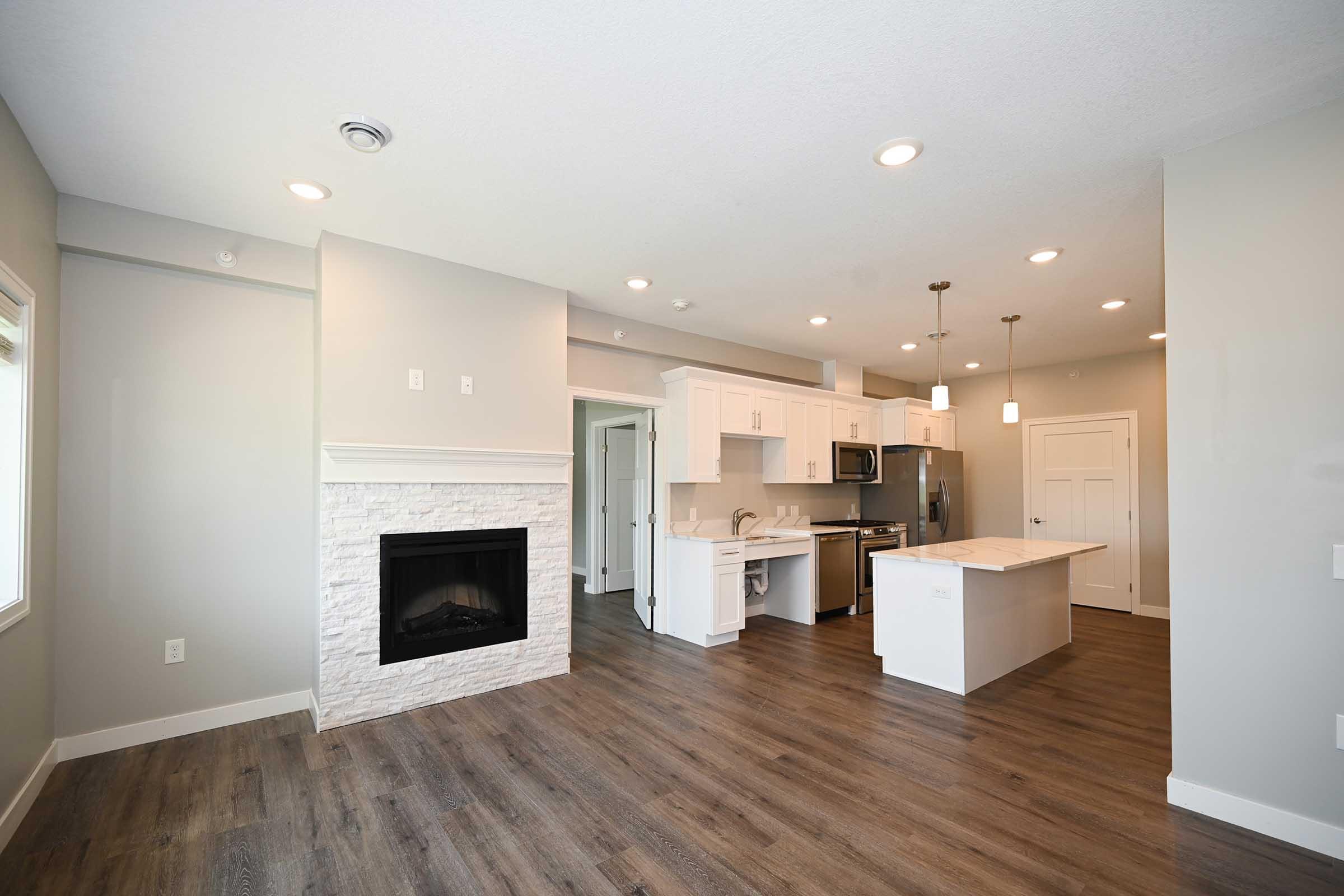 A bright and modern open-concept living space featuring hardwood floors, white cabinetry, and a cozy fireplace. The kitchen is equipped with stainless steel appliances and a large island, with natural light filling the room from multiple windows. The overall decor is neutral and inviting.