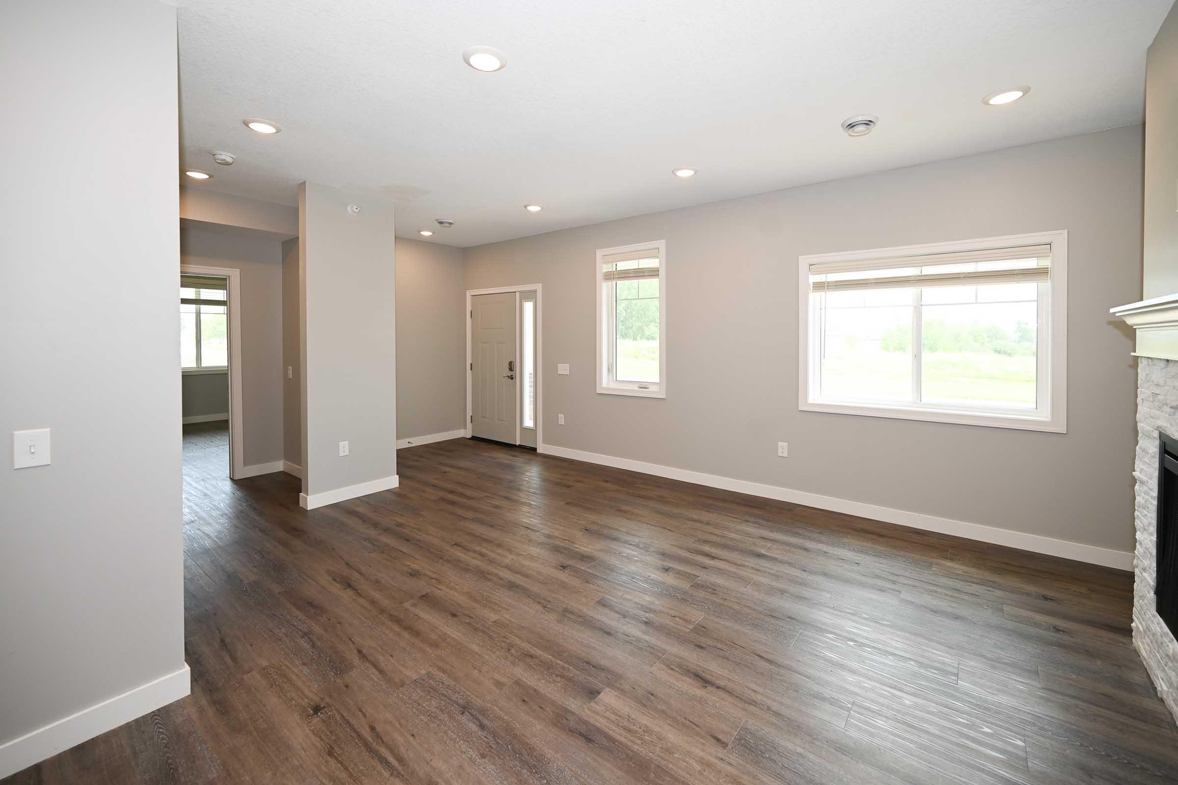 A spacious, modern living room featuring light gray walls, hardwood floors, and recessed lighting. Two windows allow natural light to fill the room, and a doorway leads to another room. The entrance door is visible, and a stone fireplace adds a cozy touch to the space.