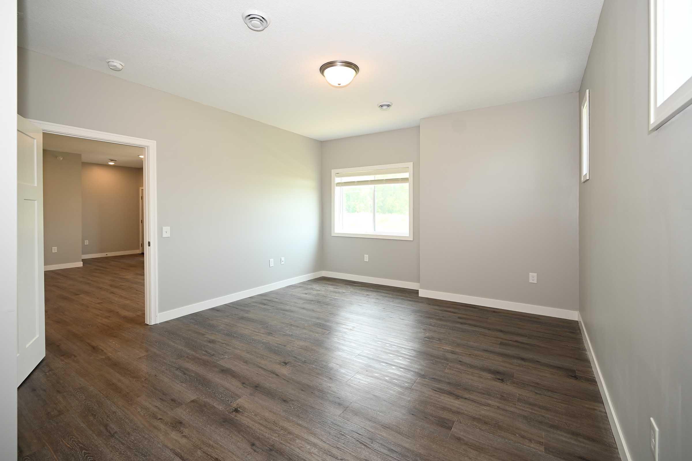 A spacious, empty room with light gray walls and a wooden floor. It features a large window providing natural light, a ceiling light fixture, and two doorways leading to adjacent spaces. The overall ambiance is modern and minimalist, emphasizing clean lines and open space.