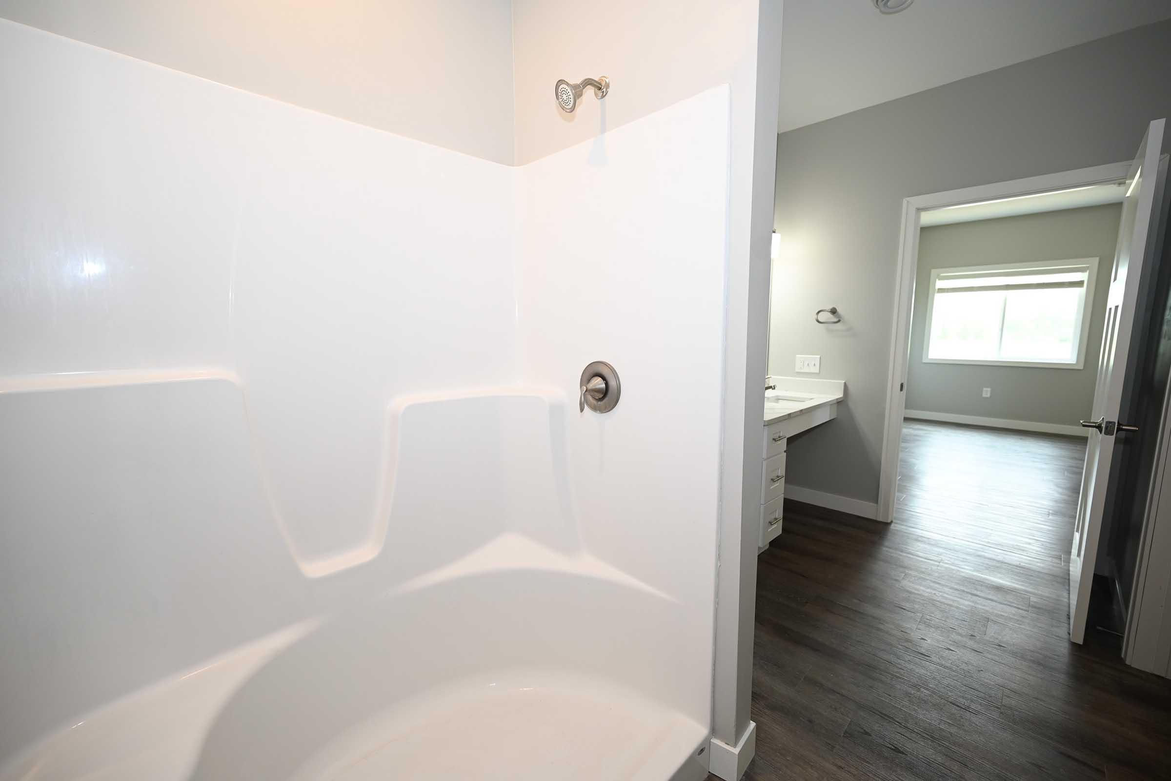 Interior view of a modern bathroom featuring a white shower stall with a single faucet, light gray walls, and a doorway leading to a spacious room with a window and bathroom vanity. The flooring is dark wood, creating a contrast with the bright shower area.