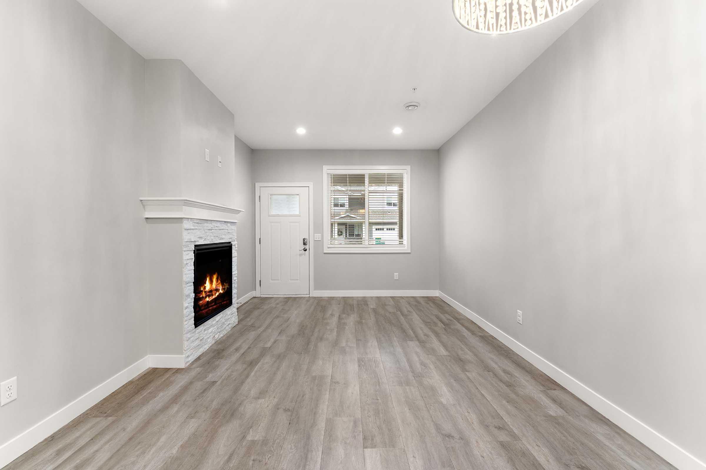 A modern, empty living room featuring light gray walls, a stylish white stone fireplace, and large windows. The floor is covered with light wooden planks, and the ceiling has a decorative light fixture. There is a front door visible, adding a welcoming touch to the space.