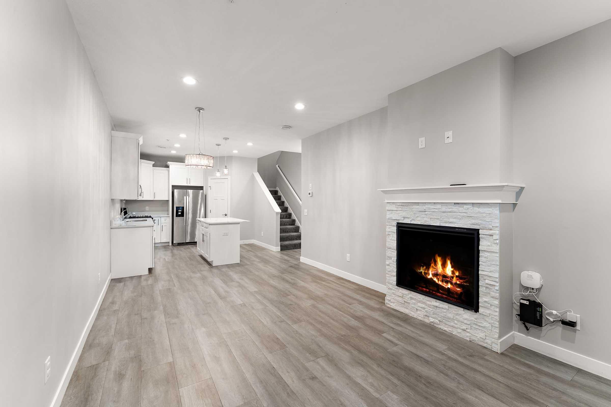 Modern living room featuring a stylish fireplace with a stone facade, light wood-like flooring, and neutral gray walls. The space includes an open kitchen area with white cabinetry and a contemporary light fixture above a central island. Stairs are visible in the background, leading to a second level.