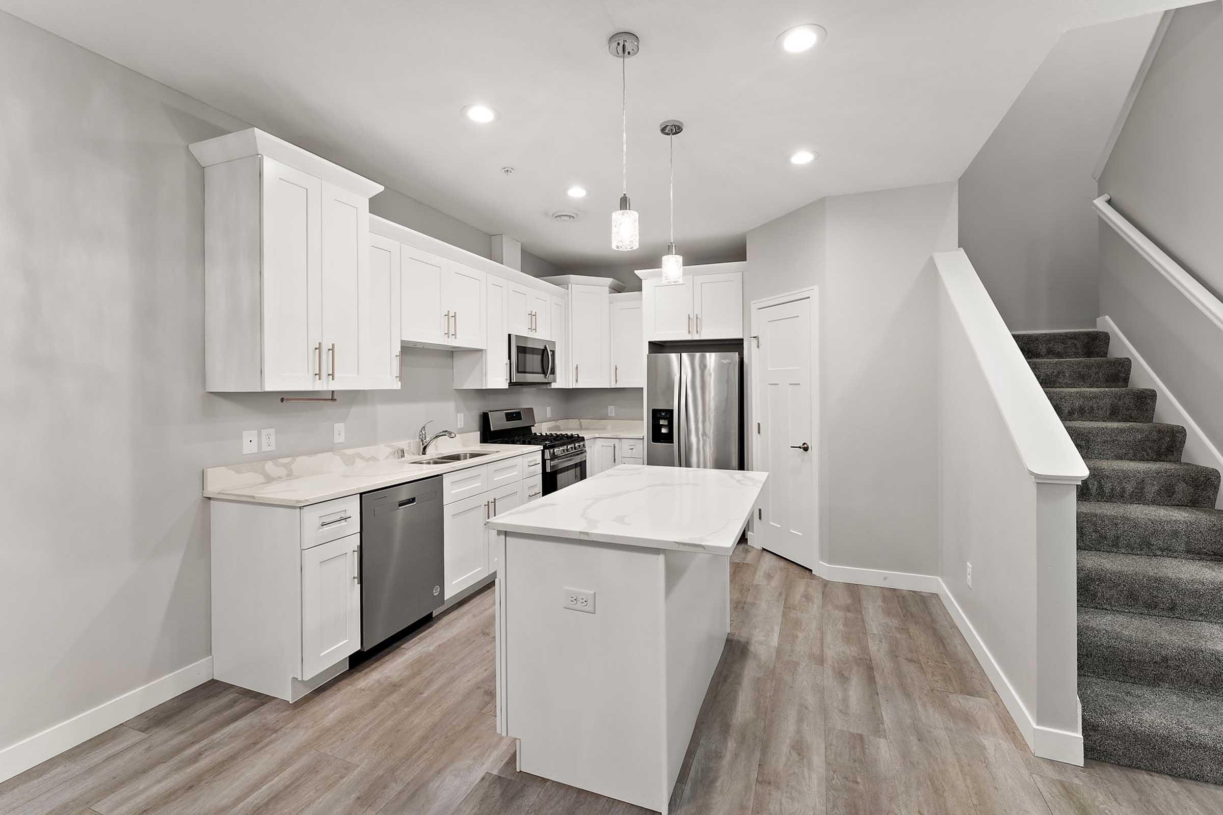 Modern kitchen with white cabinets, stainless steel appliances, and a large island. Features include a dishwasher, microwave, and a staircase leading to a second level. Bright, neutral-colored walls and warm wood flooring create an inviting atmosphere. Pendant lighting adds contemporary elegance.