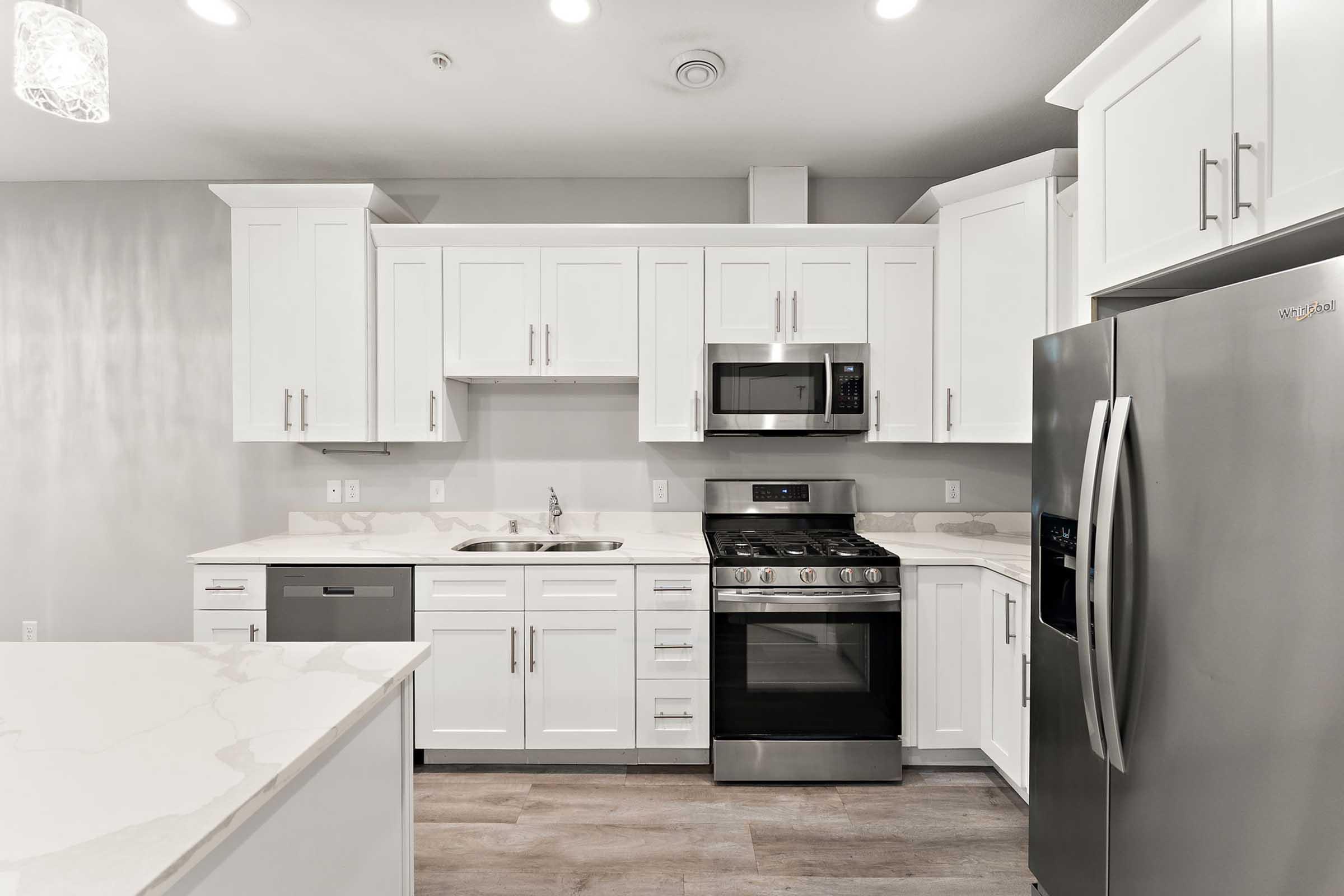 A modern kitchen featuring white cabinets, stainless steel appliances, and a marble-like countertop. The kitchen includes a refrigerator, microwave, oven, and sink, with subtle lighting and wood-like flooring creating a bright and inviting atmosphere.