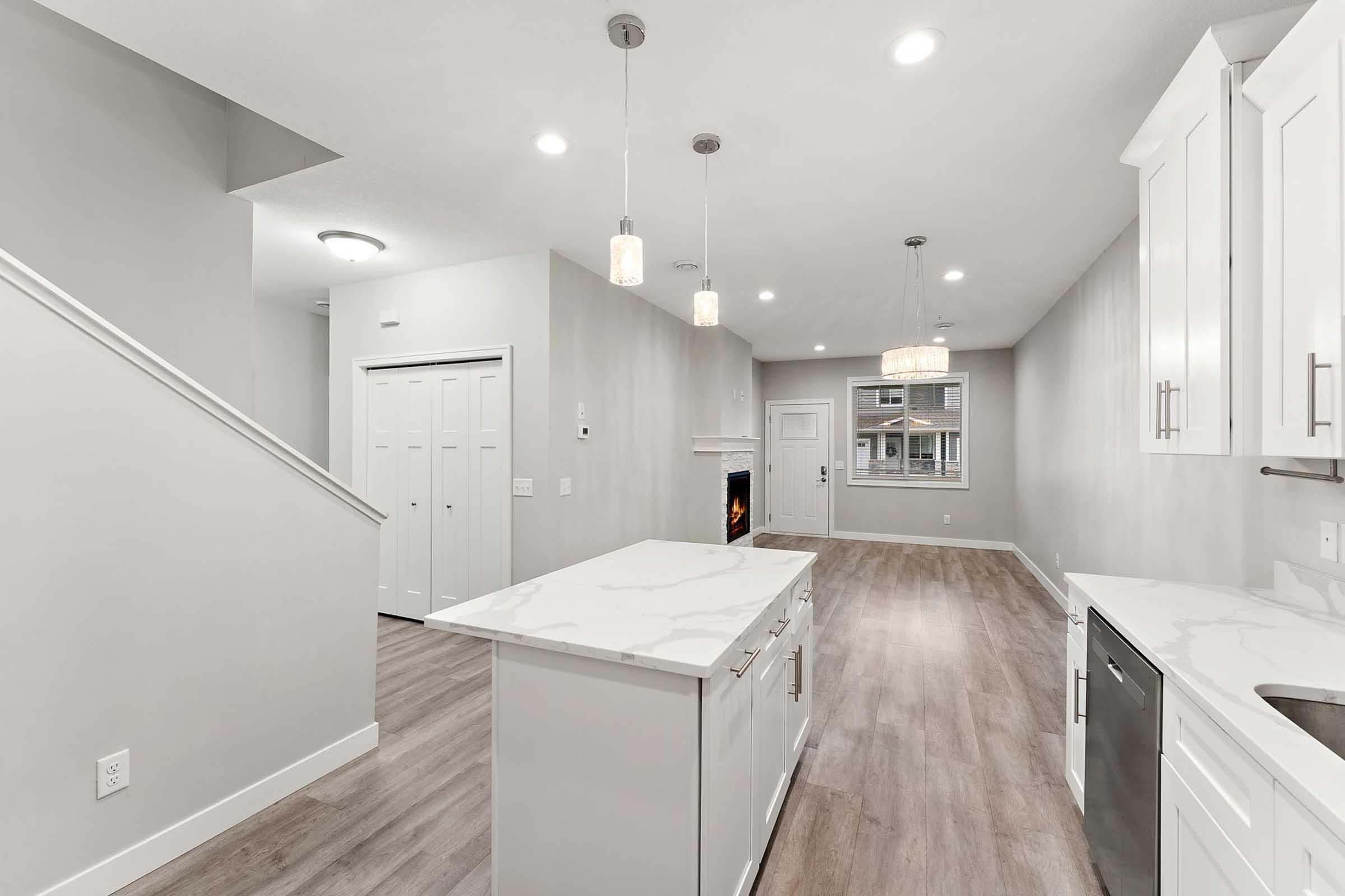 Modern open-concept kitchen and living area featuring light gray walls, hardwood flooring, and stylish pendant lighting. The kitchen includes a spacious island with a marble countertop, white cabinets, and stainless steel appliances. A fireplace is visible in the living area, creating a cozy ambiance.