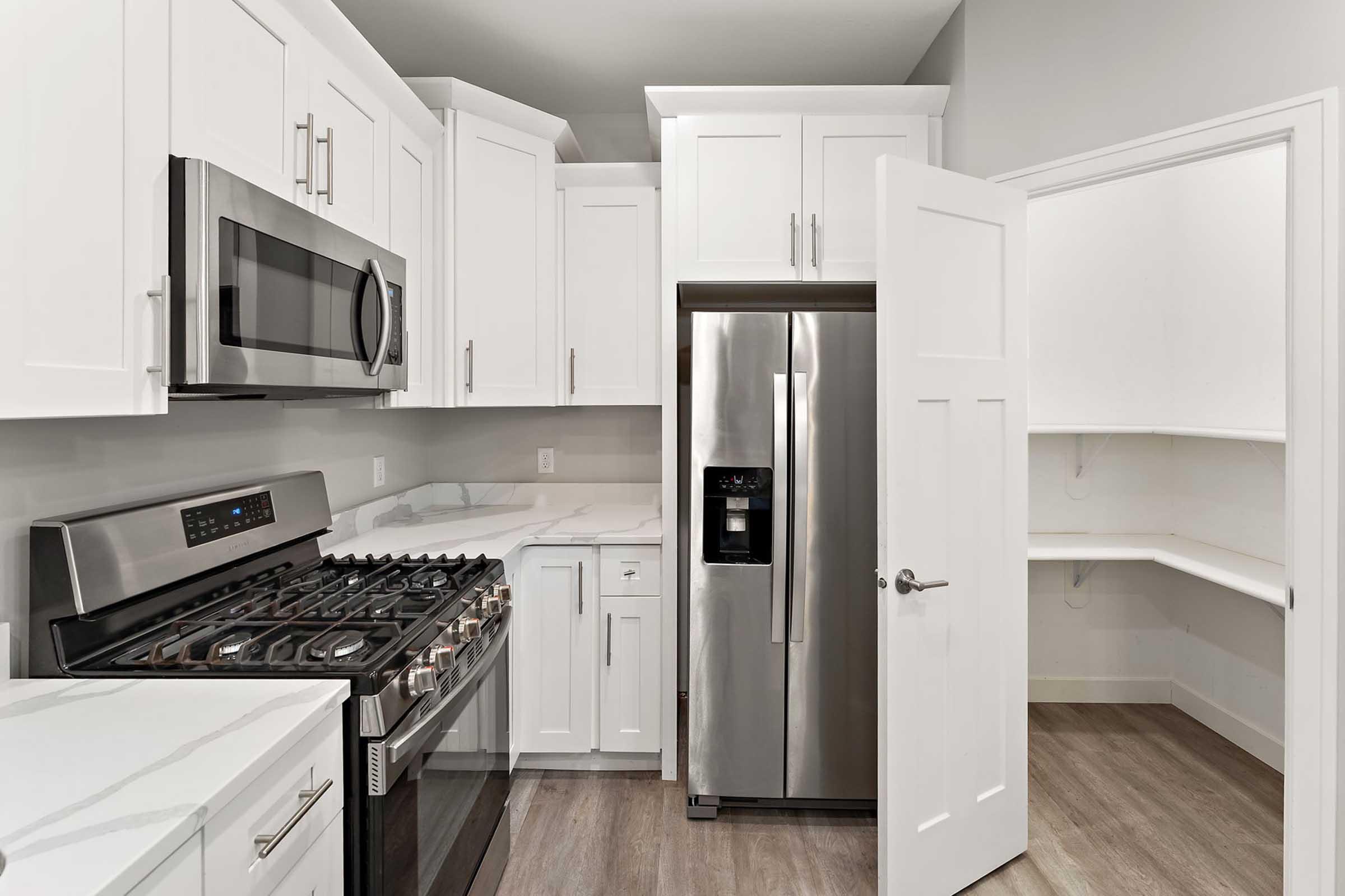 Modern kitchen with white cabinetry, stainless steel appliances including a gas stove, microwave, and refrigerator. Features a spacious layout with light-colored countertops and a pantry area with open shelving. The floor is finished with light wood laminate.