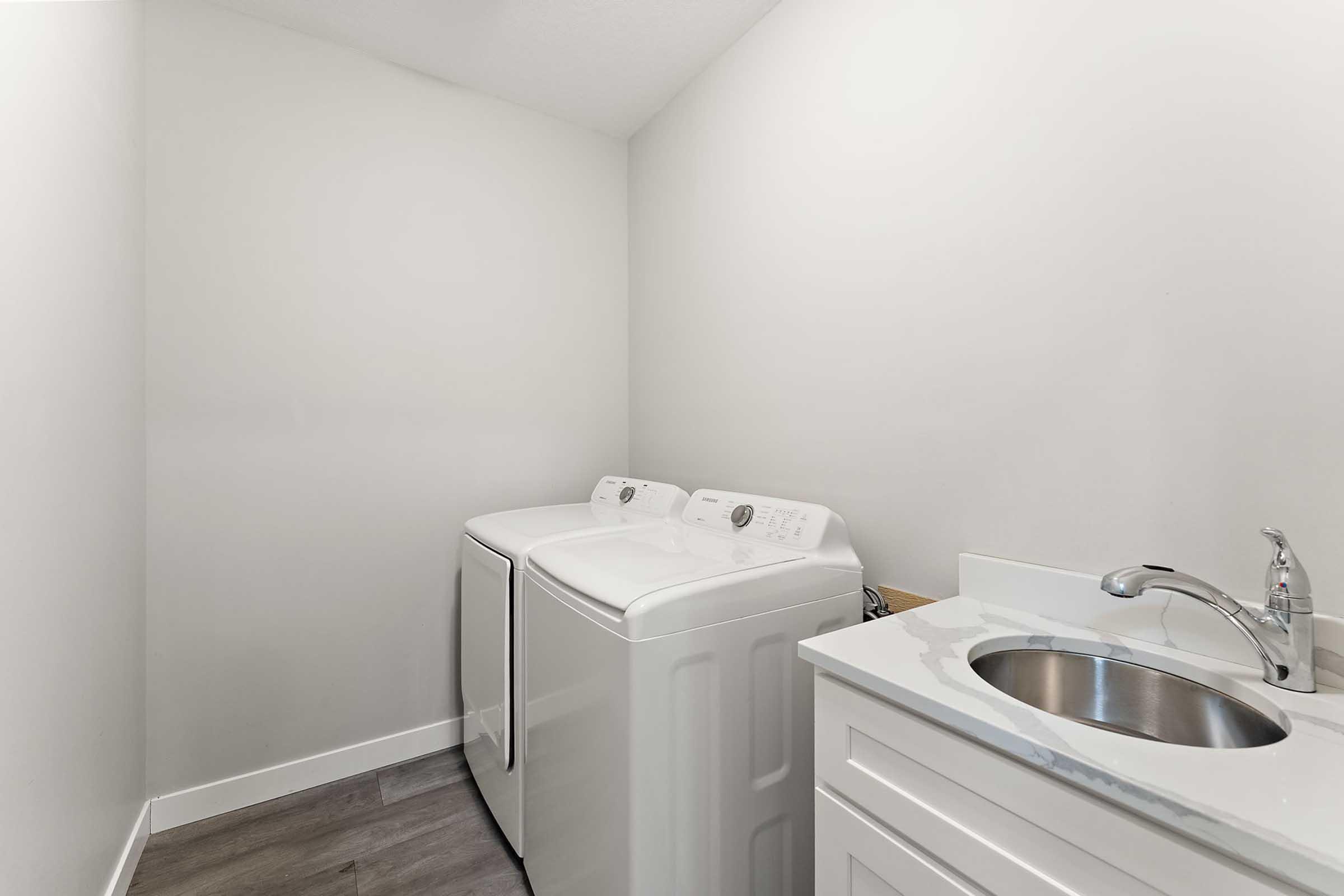 A clean, modern laundry room featuring a white washing machine and dryer side by side, with a small sink and white cabinetry. The walls are painted in a light color, and the flooring is a neutral gray, creating a bright and organized space for laundry tasks.