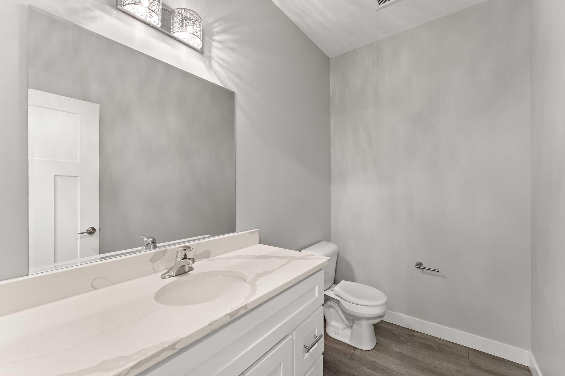 A modern bathroom featuring a light gray color palette. It has a white vanity with a marble countertop and a sink, a wall-mounted mirror, and a toilet. Soft lighting fixtures are installed above the mirror, and the flooring is composed of light-colored tiles. The overall design is clean and minimalist.